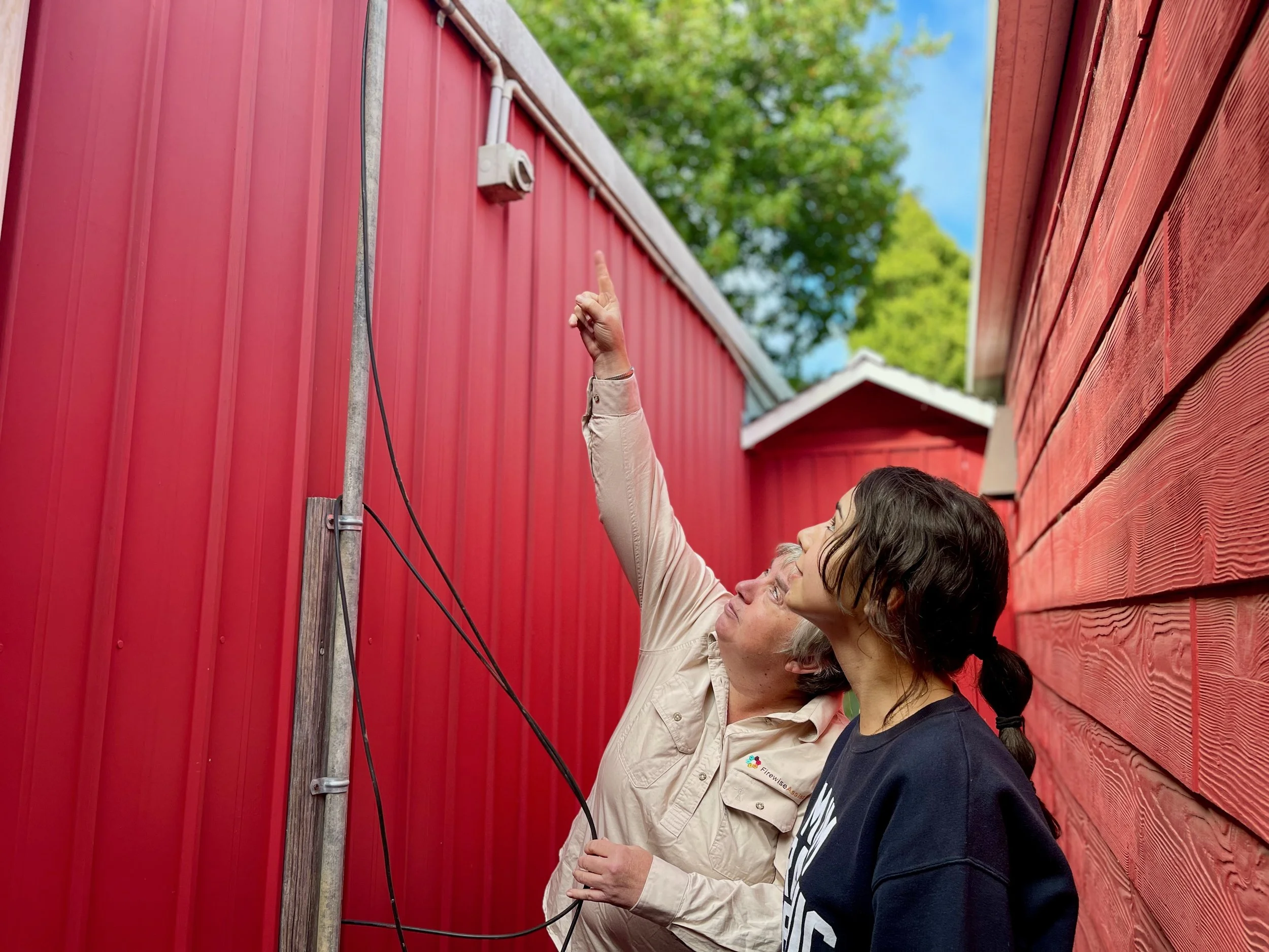 Two people examining an outdoor security camera mounted on a red wooden fence between two red buildings, with green trees and a blue sky in the background.