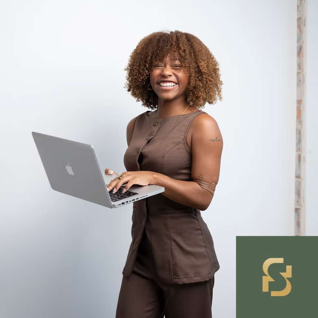 Woman with curly hair smiling and holding a MacBook laptop, standing against a white wall. Logo in corner.