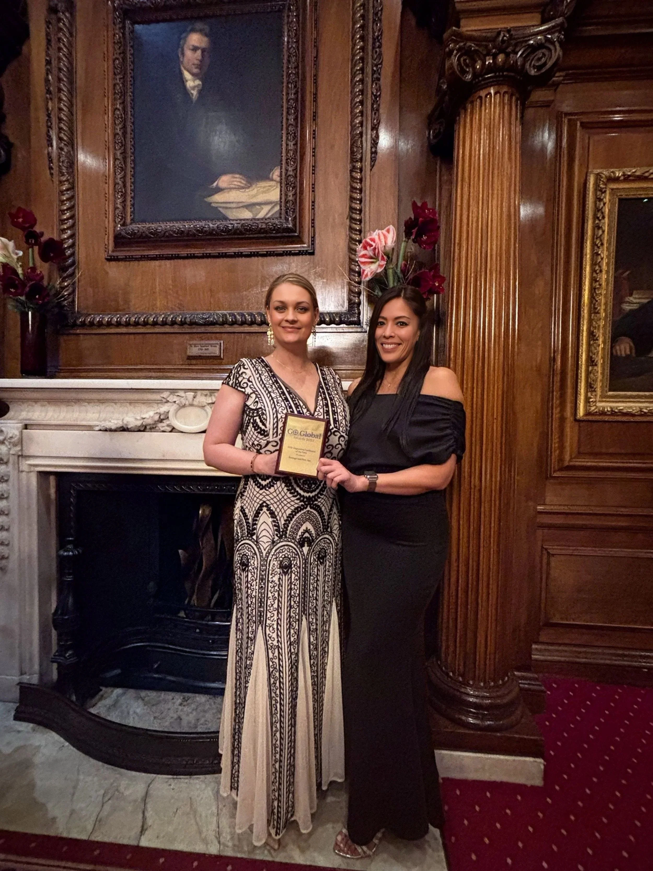 Two women standing in a room with wood-paneled walls and a fireplace, holding an award or certificate. One woman is wearing a patterned dress, and the other is dressed in black. There is a large portrait of a man hanging on the wall behind them, and floral arrangements near the portrait.