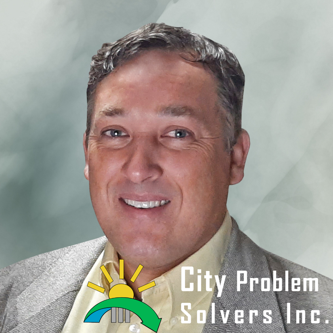 A smiling middle-aged man with short brown hair, wearing a light-colored shirt and gray blazer, against a muted background with a logo for 'City Problem Solvers Inc.' featuring a sun, bridge, and arrow.