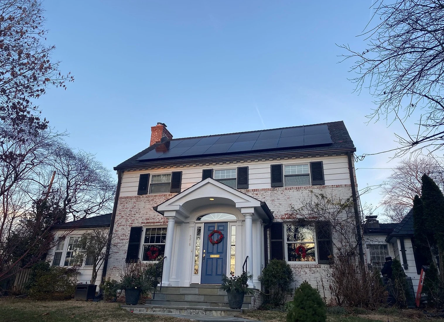 Two-story colonial-style house with solar panels on the roof, a blue front door, and Christmas wreaths on the windows.