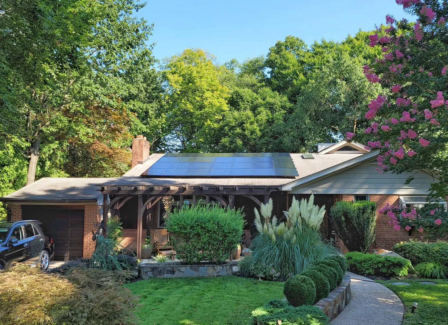 House with solar panels on the roof, surrounded by trees and a well-maintained garden, featuring a driveway with a parked car.