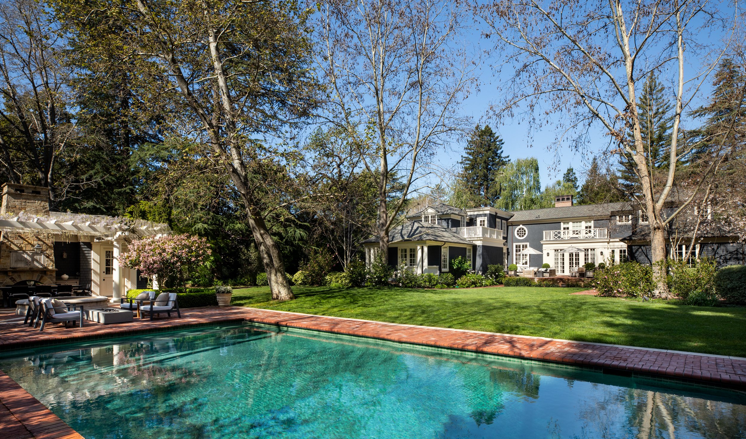 A backyard with a swimming pool, patio furniture, large trees, and a multi-story house with white trim and black siding under a clear blue sky.