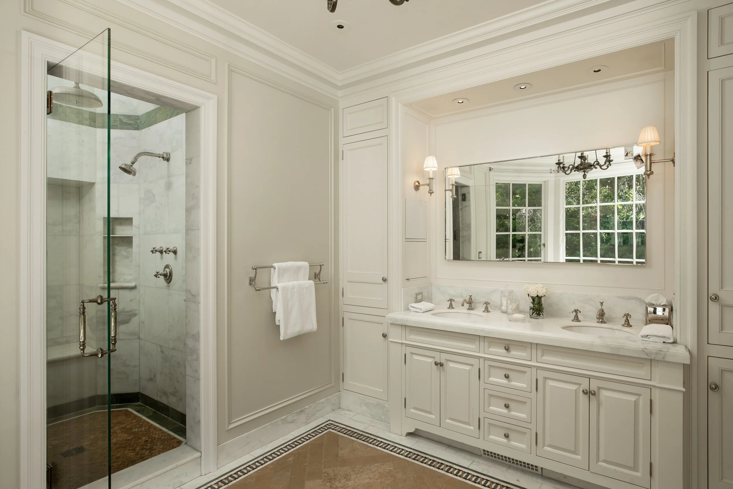 A luxurious bathroom with a glass-enclosed shower, white cabinetry, marble countertops, holiday lights, and large windows with greenery outside.