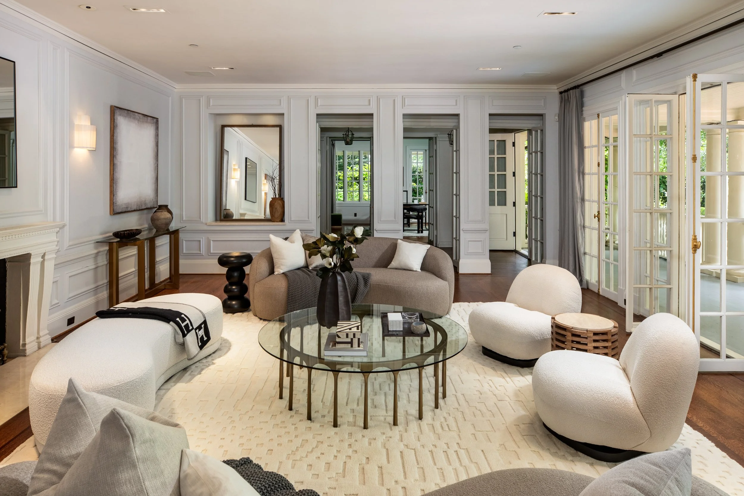 Living room with white walls, a beige sectional sofa with white pillows, a glass coffee table with books and decorative objects, and large windows with curtains allowing natural light in.