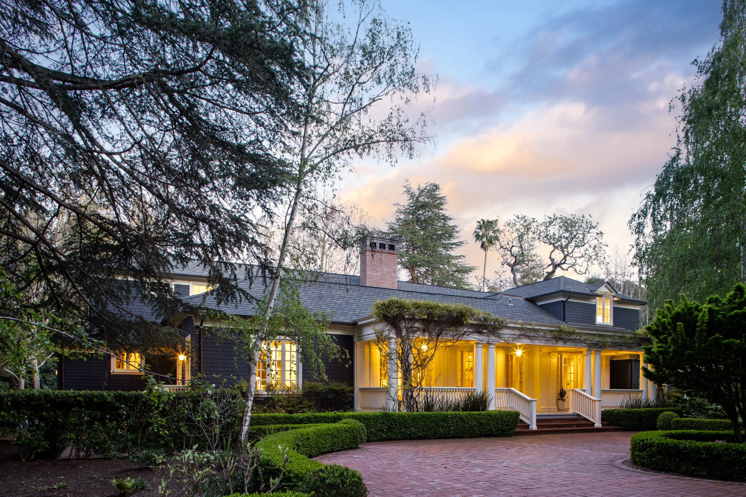 A large house with a porch illuminated at dusk, surrounded by trees and a well-maintained garden with bushes and a brick pathway.