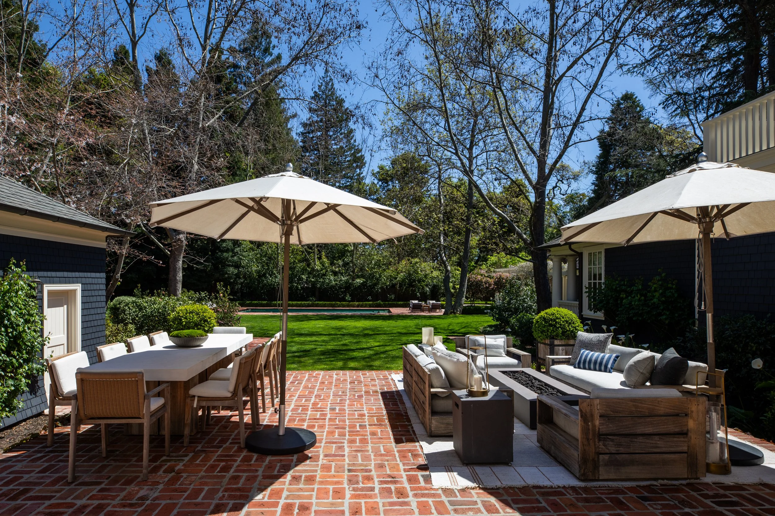 A backyard patio with outdoor furniture, umbrellas, and lush greenery, including trees and a lawn, under a clear blue sky.