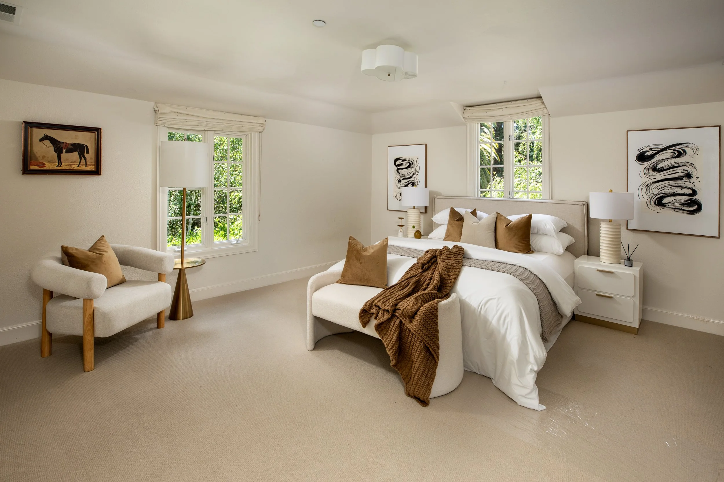 A bedroom with a large bed, beige headboard, and white bedding decorated with brown pillows and a brown throw. There are two white nightstands with white lamps on each side of the bed, and a white armchair with a beige pillow near the left window. Th
