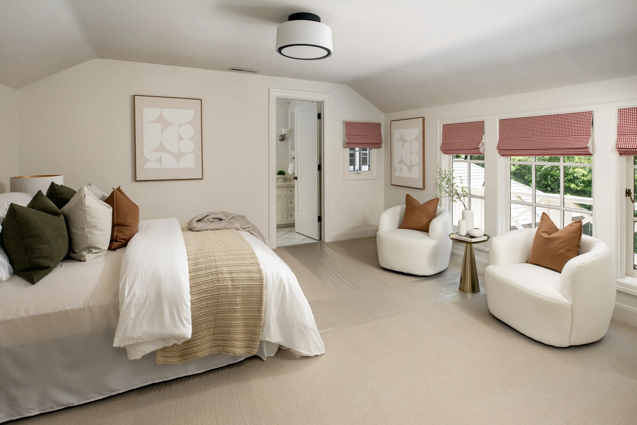 A bedroom with a bed on the left, decorated with multiple pillows, a blanket, and a bedside lamp. There are two white armchairs with brown pillows near the large window with red checkered curtains. The room has light-colored walls, art on the wall, a