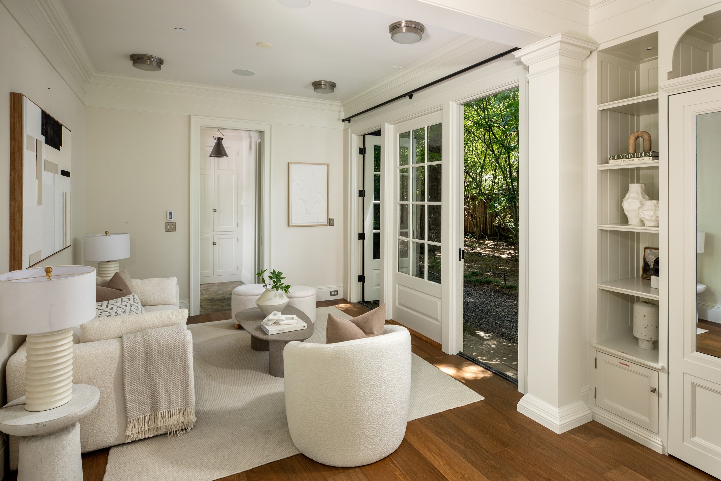 Living room with white furniture, a coffee table, and a view of a garden through French doors.