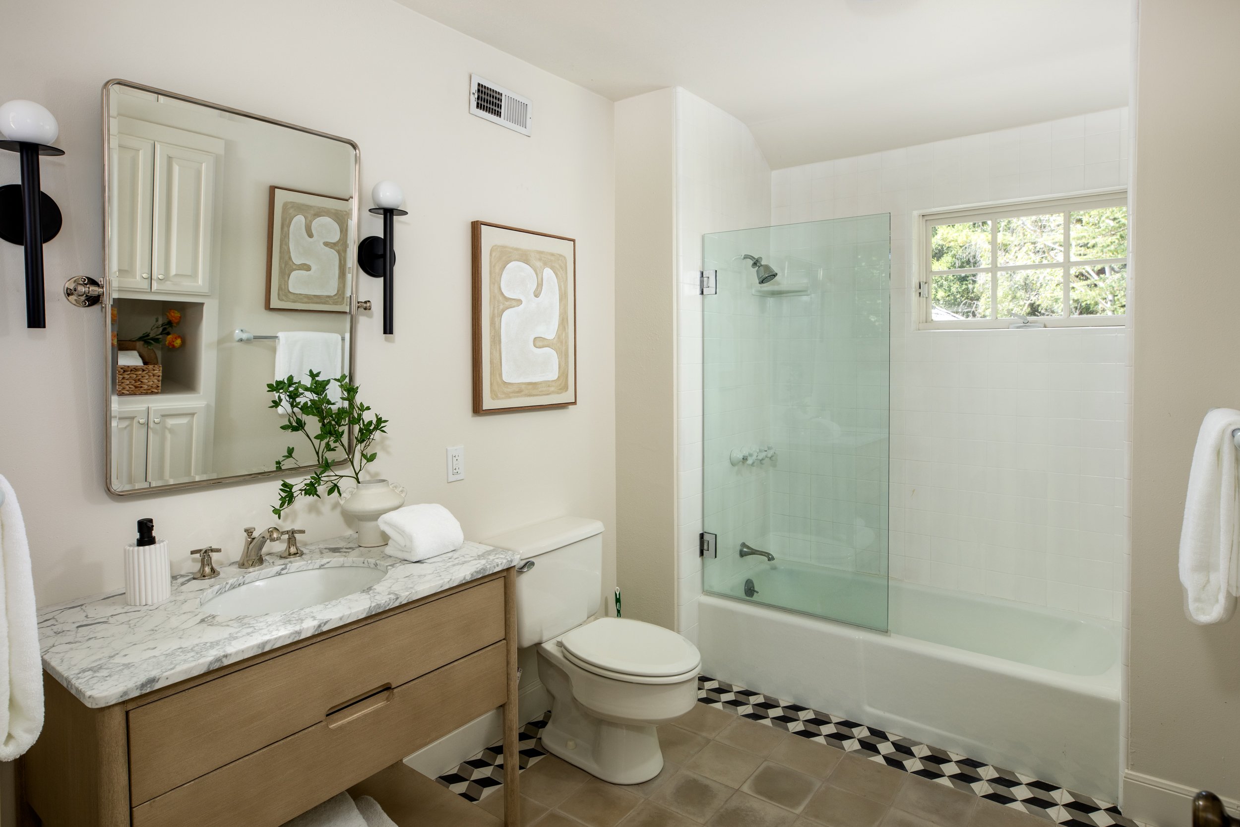 A bathroom with a white vanity, marble countertop, and a mirror. There is a toilet next to the vanity, and a glass-enclosed shower with a window showing greenery outside.