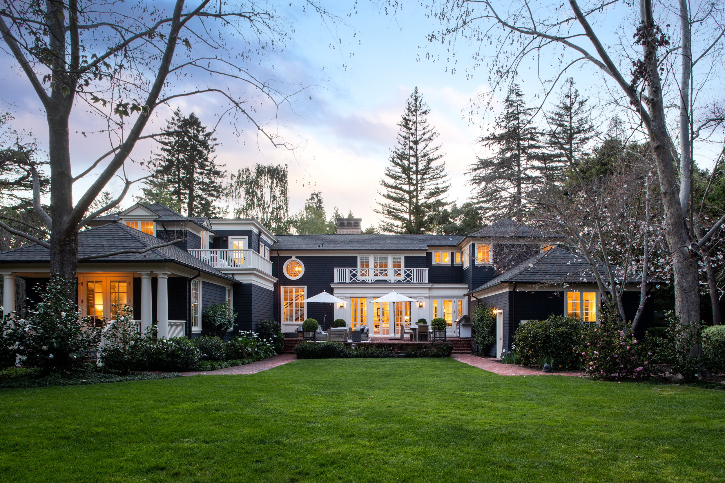 A large dark blue house with multiple stories, large windows with lights on, surrounded by trees and a well-manicured lawn in the evening.