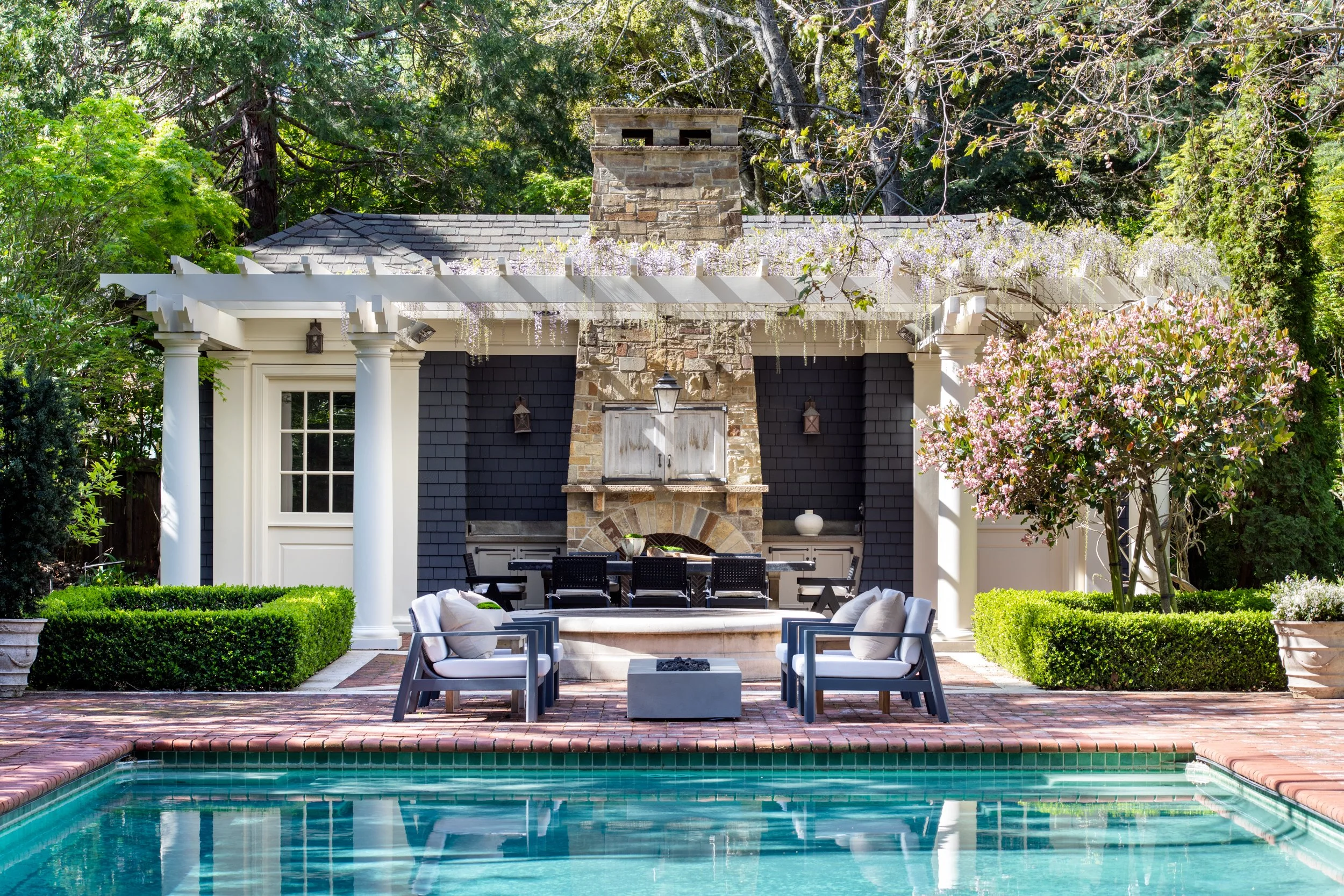 Residential backyard with a swimming pool, outdoor furniture, a stone chimney, and a pergola decorated with hanging purple flowers, surrounded by lush green trees and bushes.