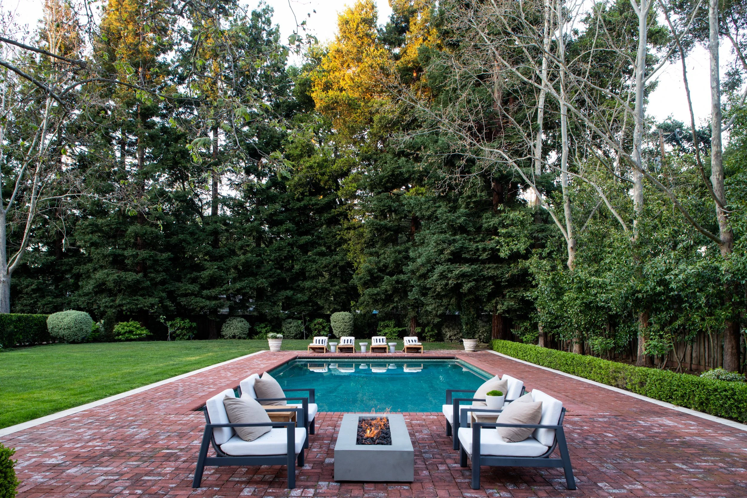 Backyard patio with an in-ground pool, outdoor furniture, a fire pit, and surrounded by well-maintained grass, shrubs, and tall trees.