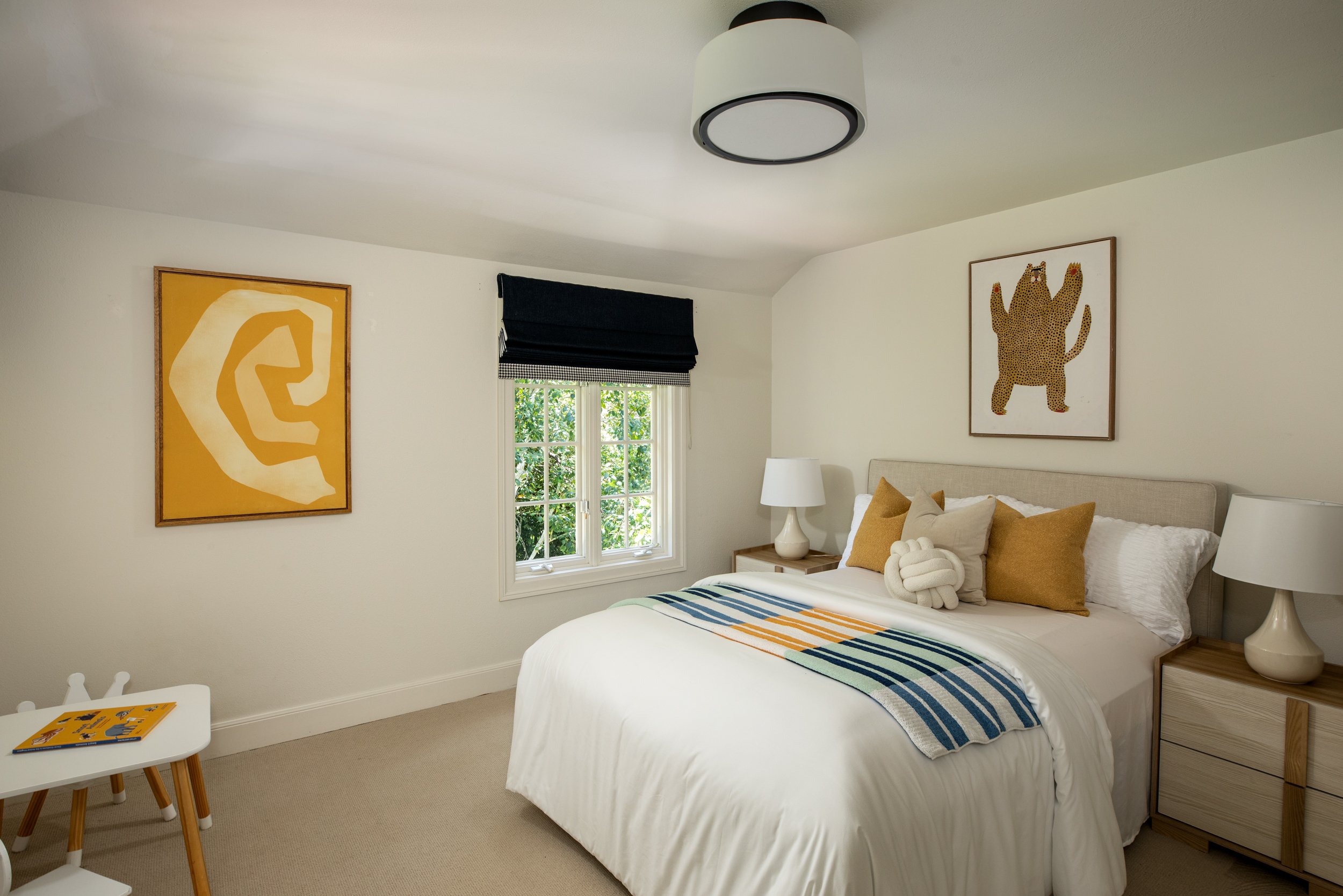 A neatly made bedroom with a white bed, beige headboard, and decorative pillows, flanked by matching nightstands with white lamps. Two framed animal-themed artworks decorate white walls, a window with black Roman shade, and a striped throw blanket on