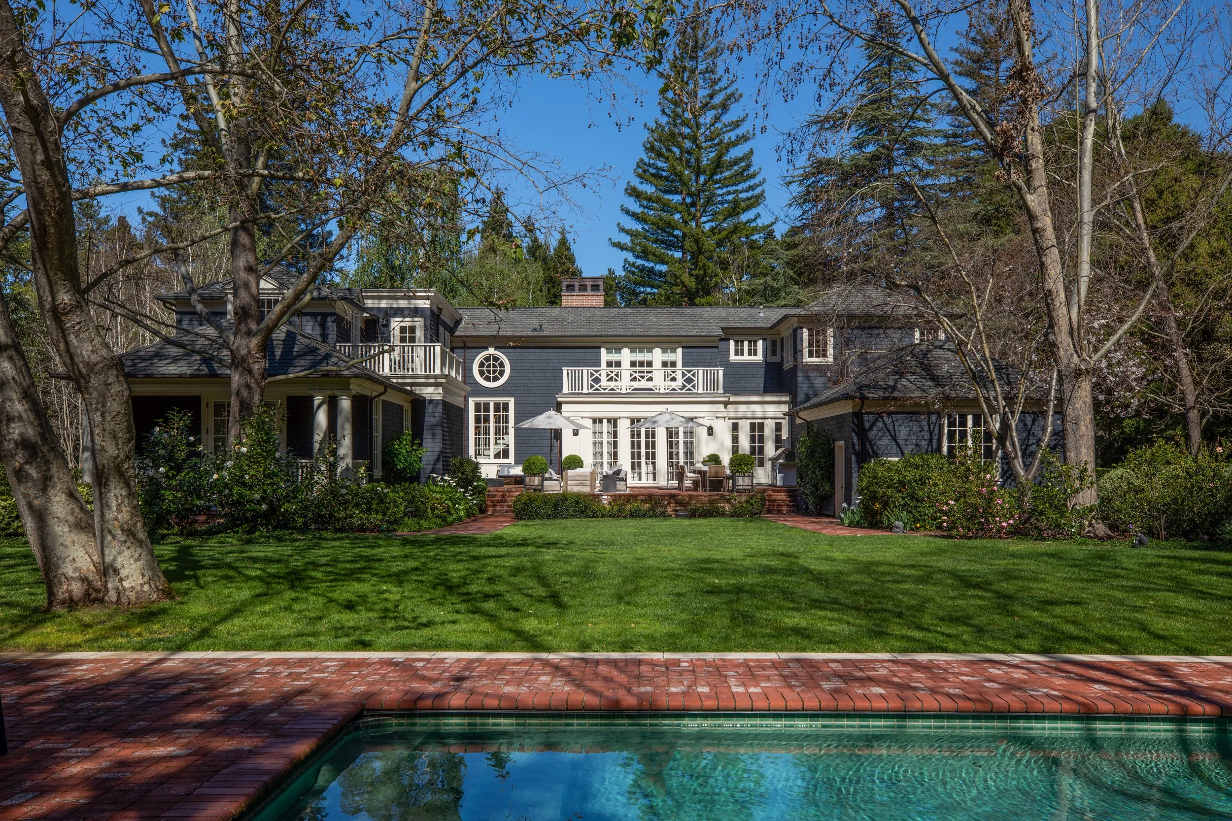 Backyard view of a large house with a patio, umbrellas, and outdoor furniture, surrounded by green grass, trees, and shrubs, with a swimming pool in the foreground.