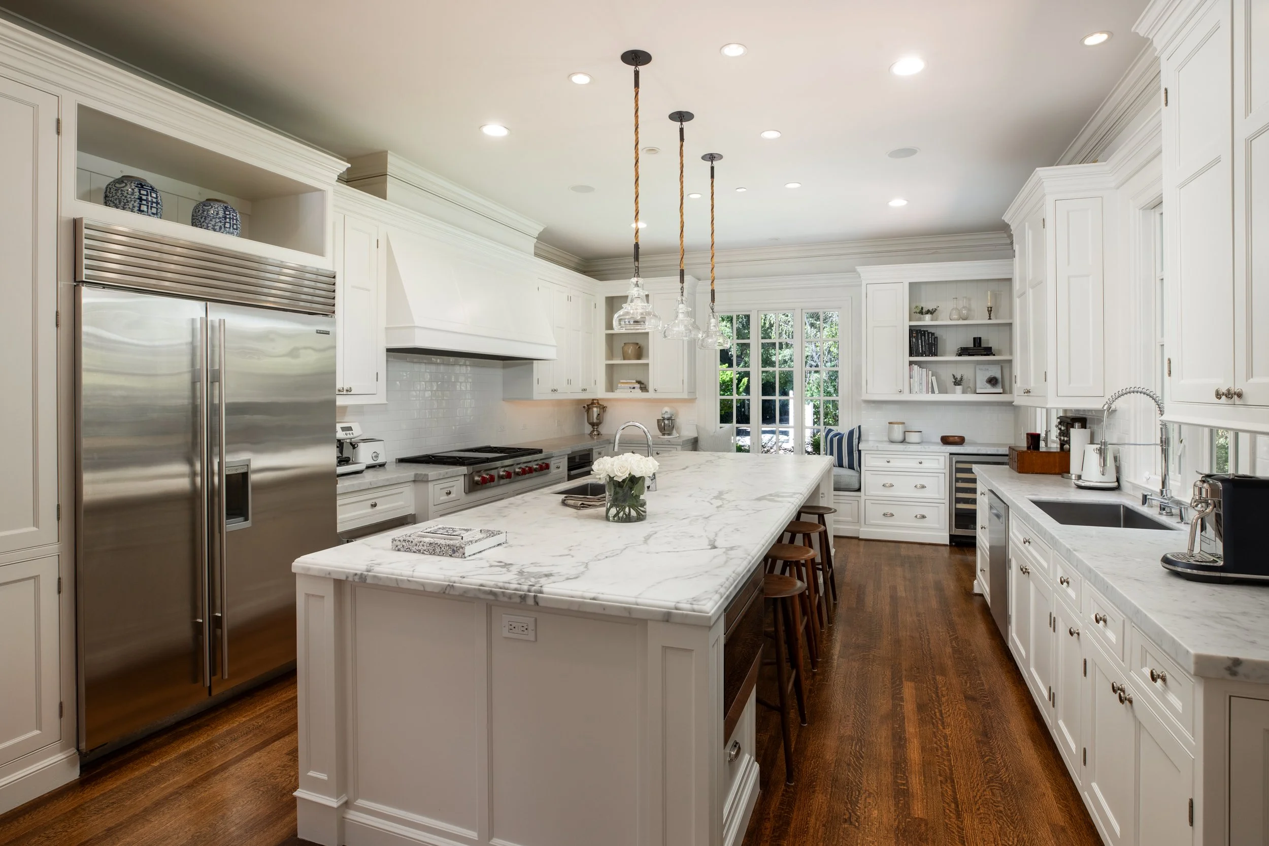 A spacious kitchen with white cabinets, marble countertops, and a large island. The kitchen features stainless steel appliances, including a refrigerator and a professional stove with a hood. There are pendant lights hanging over the island, and a la