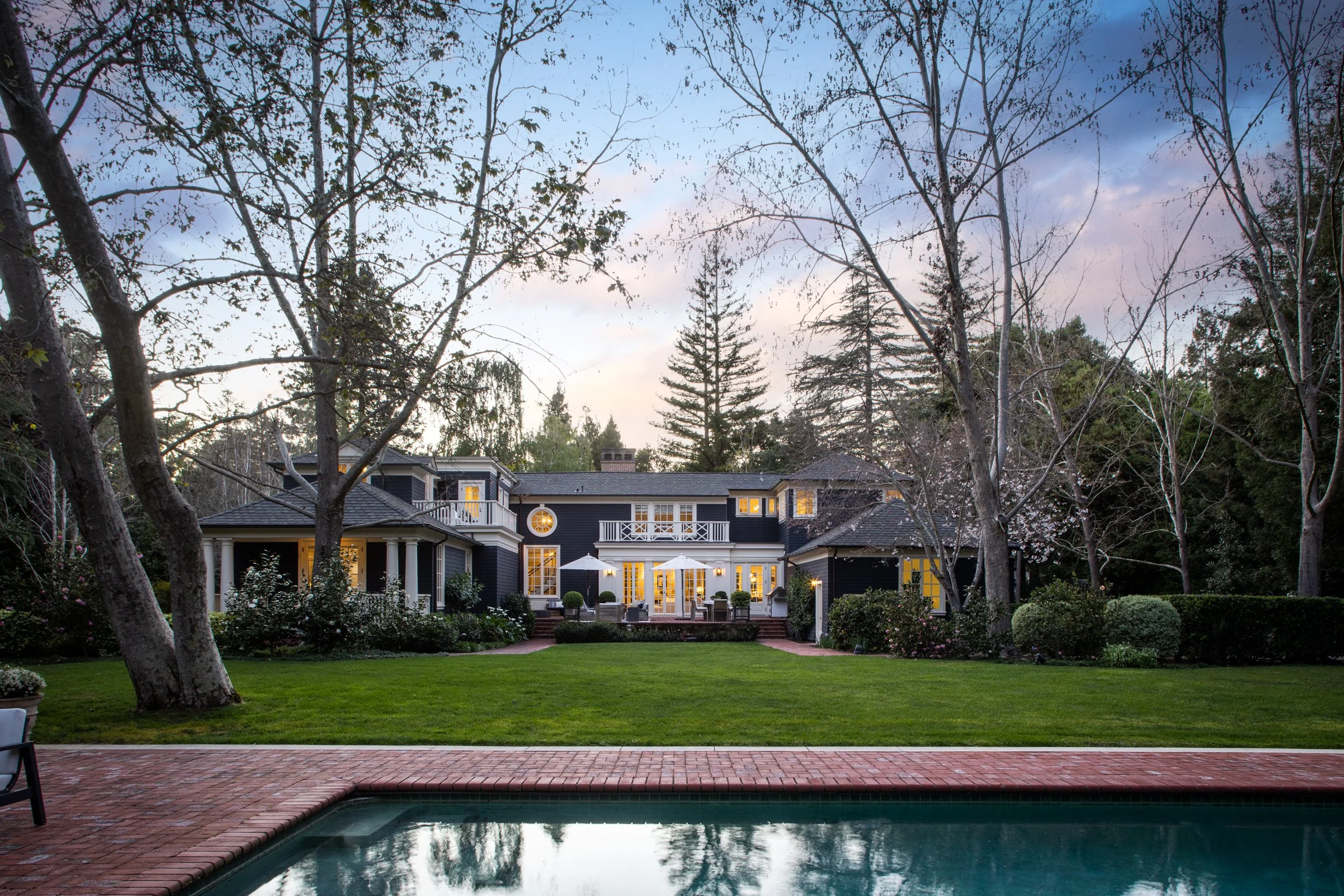 Backyard view of a large, dark-colored house with multiple windows and a porch, surrounded by tall trees and a well-maintained lawn, with a swimming pool in the foreground reflecting the sky and trees, during dusk.