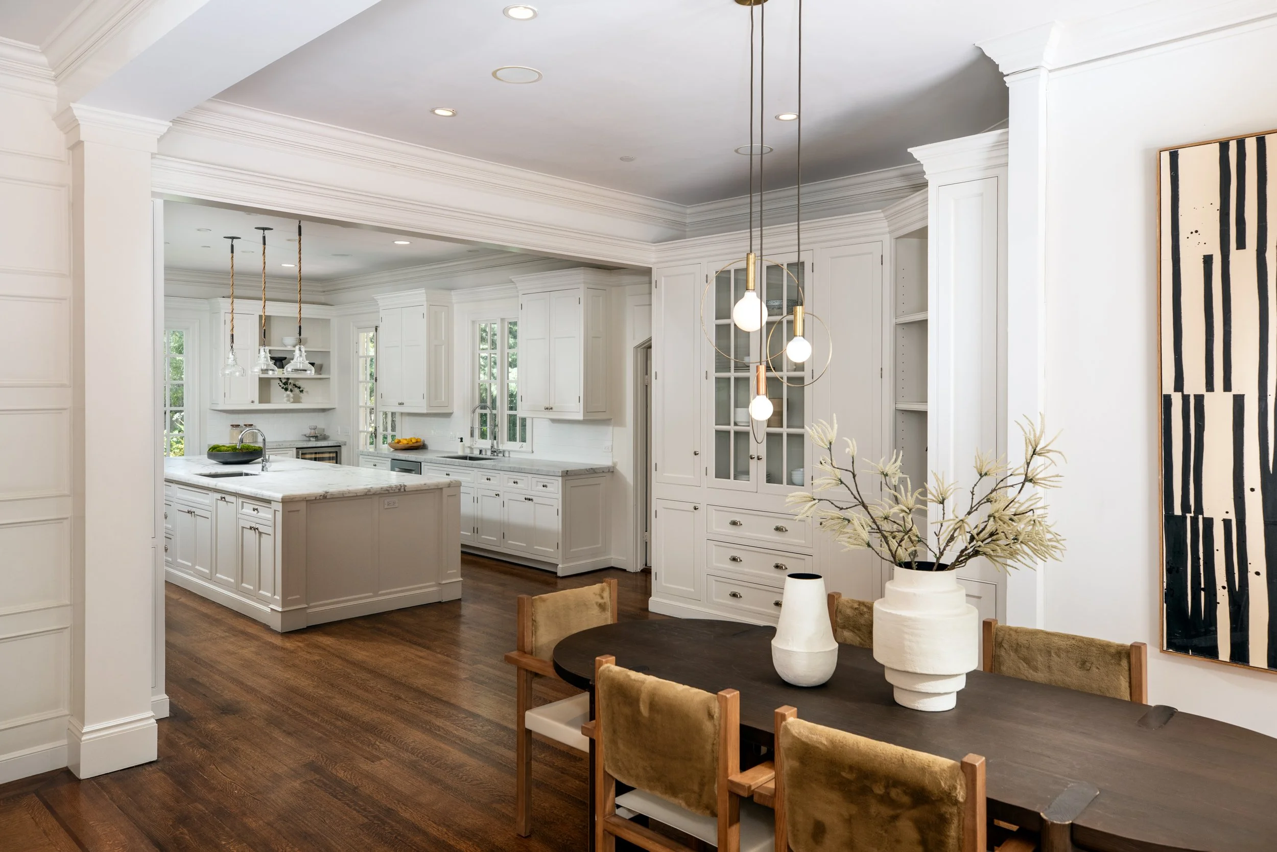 A spacious kitchen with white cabinetry, a large kitchen island with a marble countertop, pendant lights, and large windows. There is a dining table with chairs in the foreground and a piece of modern black and white art on the wall.