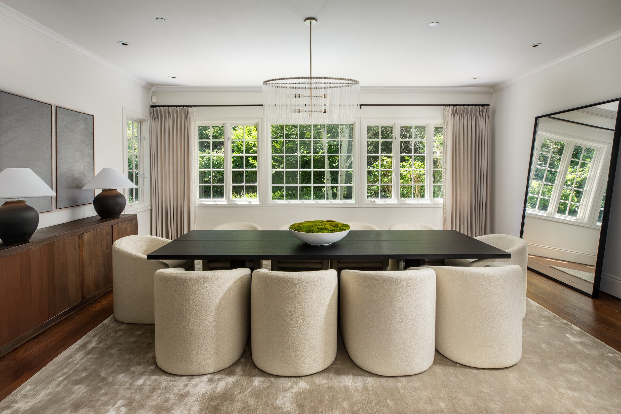 A modern living room with large windows, a round chandelier, a dark wood dining table with cream upholstered chairs, and a large leaning mirror.