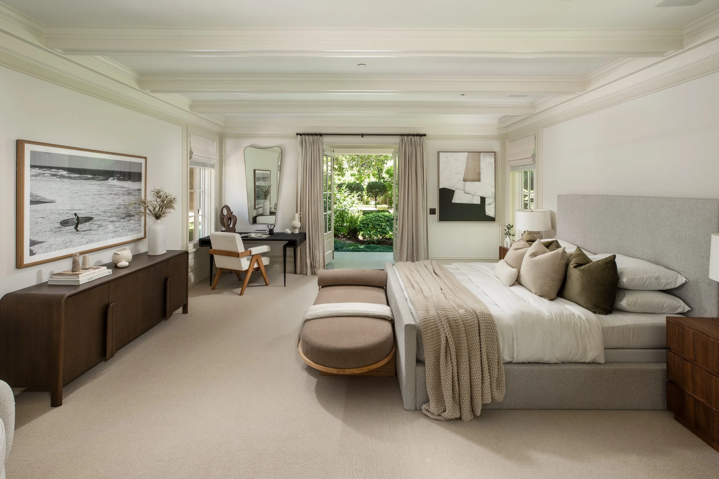 A bright, modern bedroom with a large bed, decorative pillows, a beige blanket, a gray upholstered headboard, and a bench at the foot of the bed. A wooden dresser with art and decor is against the left wall. A workspace with a black desk, white chair
