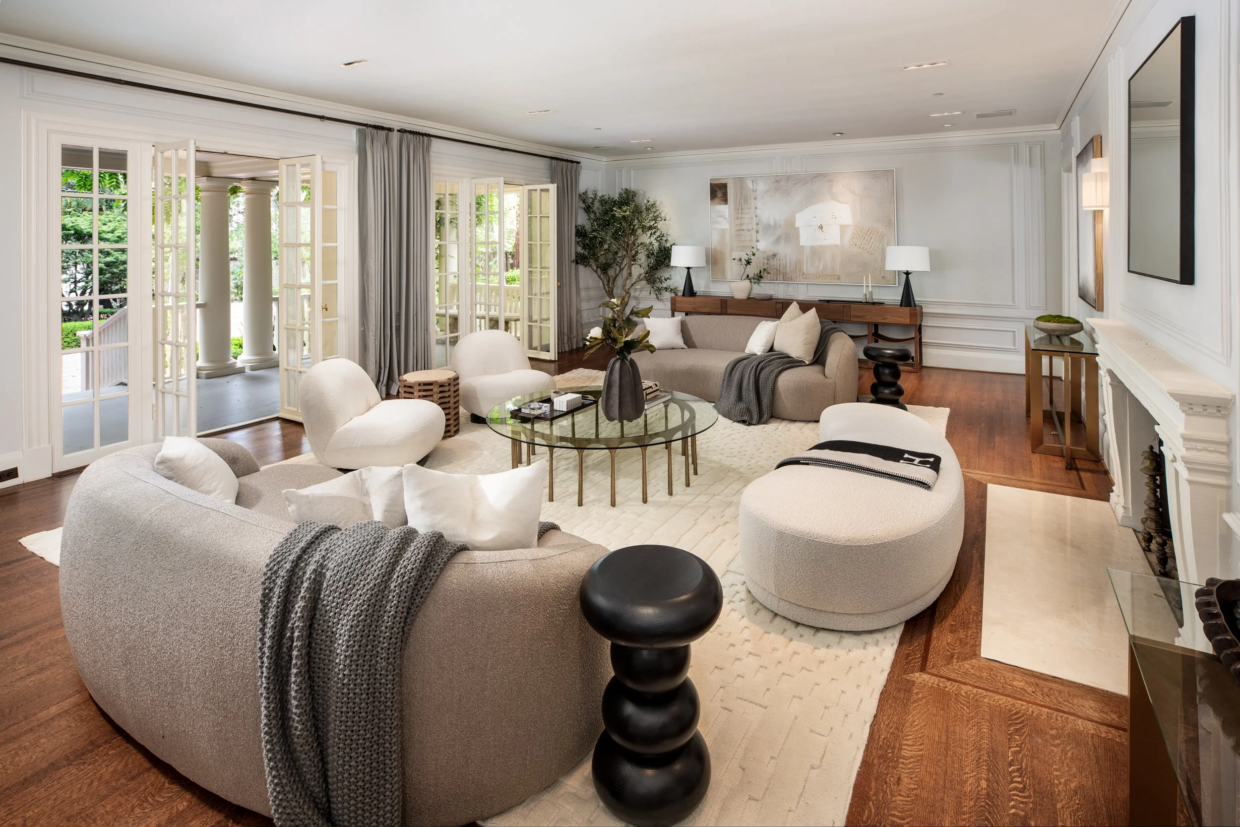 Spacious living room with white walls, large windows, neutral-colored furniture, and hardwood floors. Decor includes plants, artwork, and lamps.