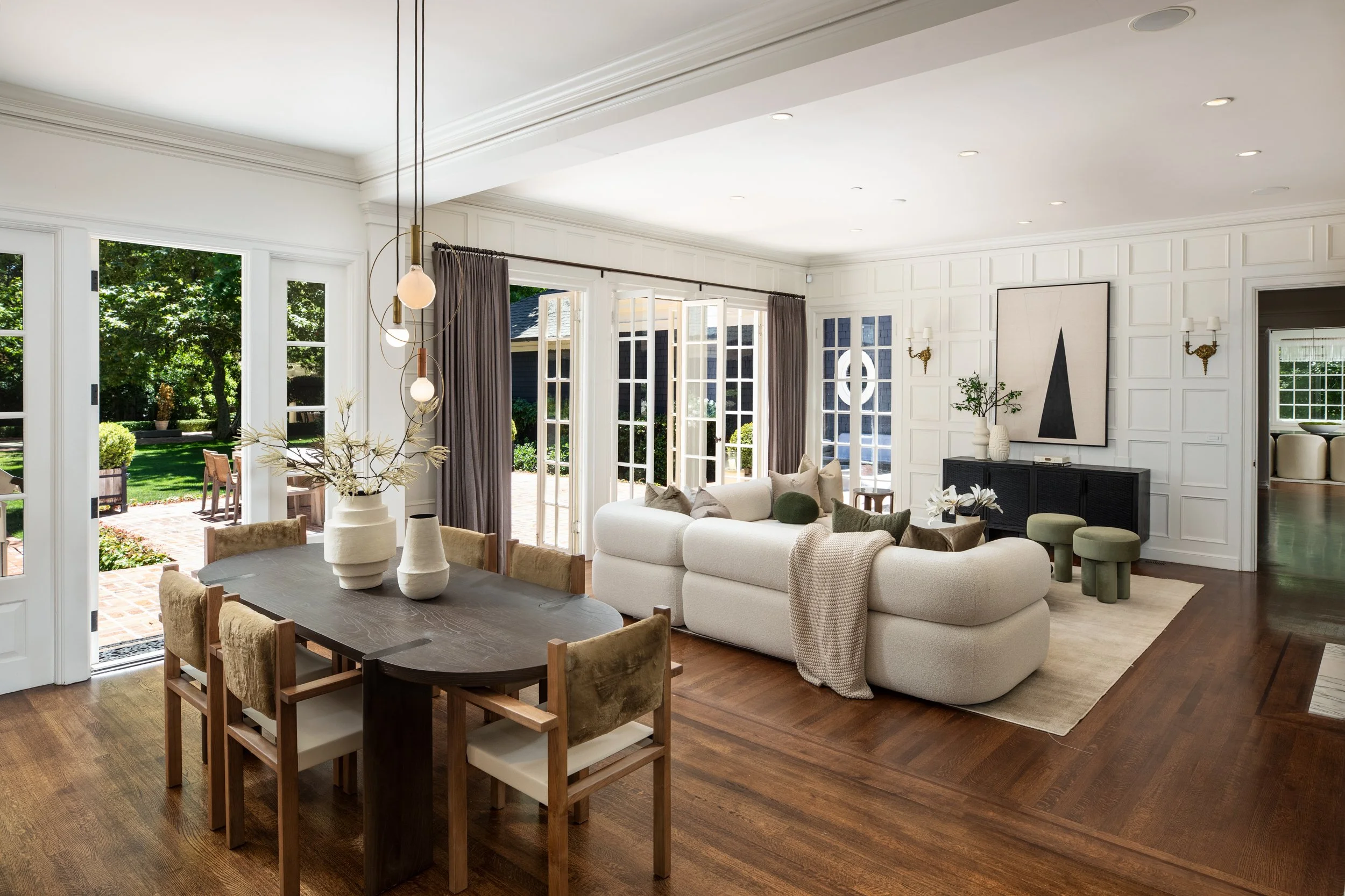 Bright and spacious living/dining room with large windows, open door to patio, hanging pendant lights, white walls with decorative molding, modern art, and various seating options.