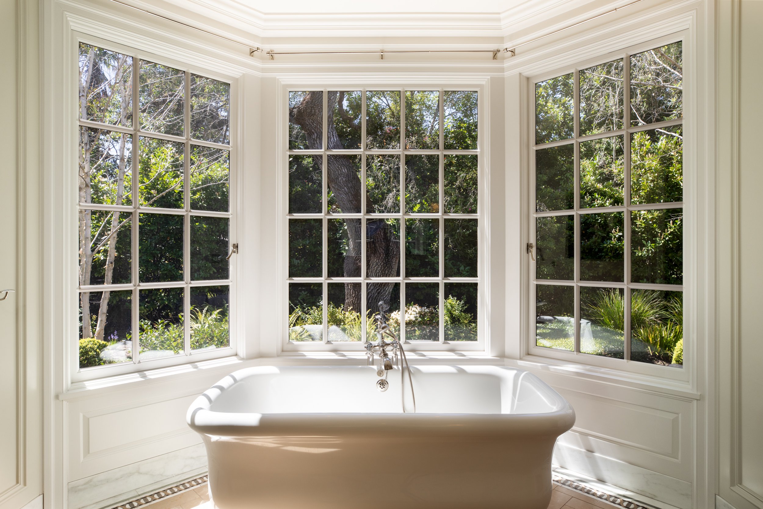 A view from inside a bathtub looking out a large bay window with a garden and trees outside.