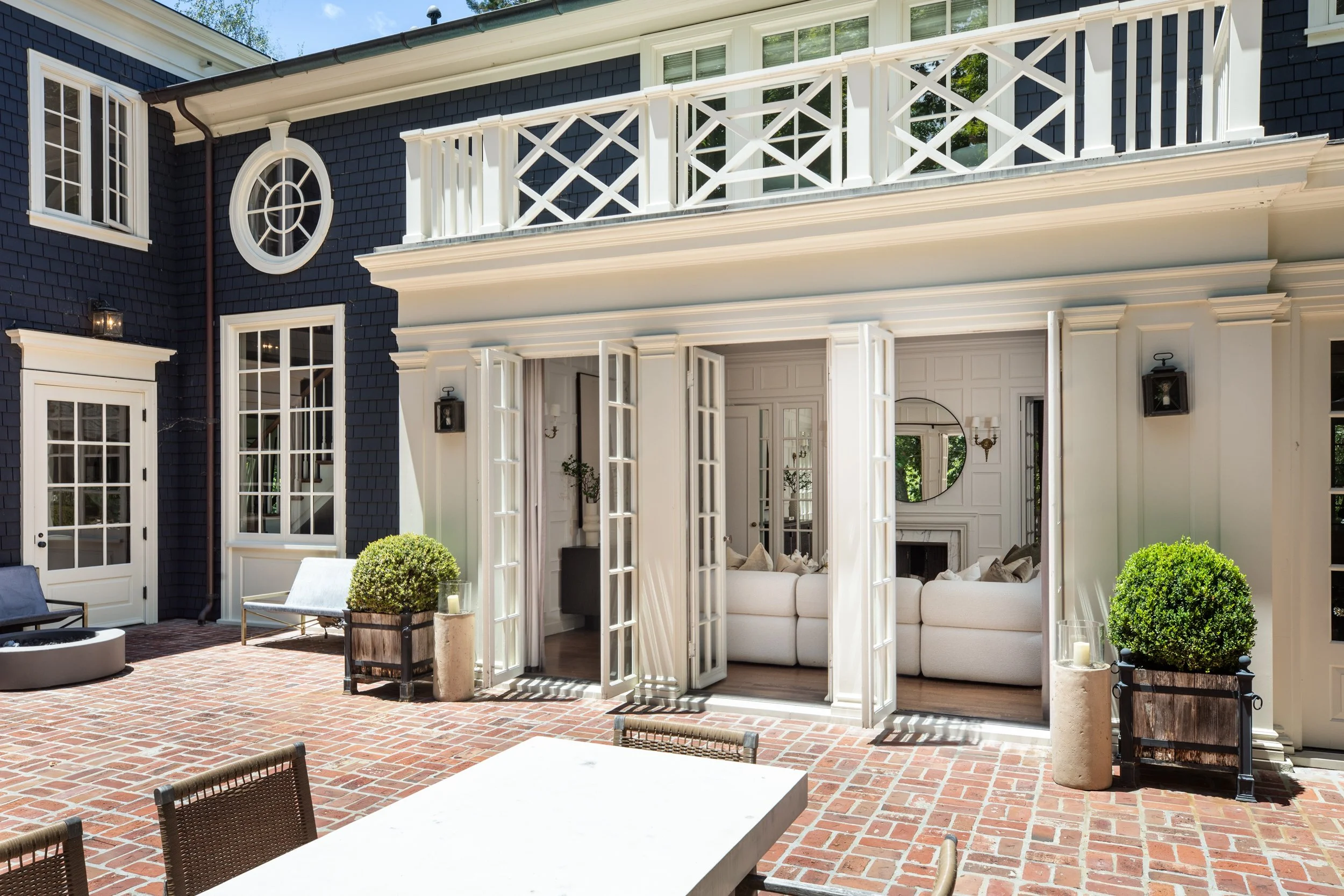Exterior view of a house with black shingle siding, white trim, multiple windows, and a white balcony. There is an open door leading to a porch with patio furniture and potted plants on a brick patio.