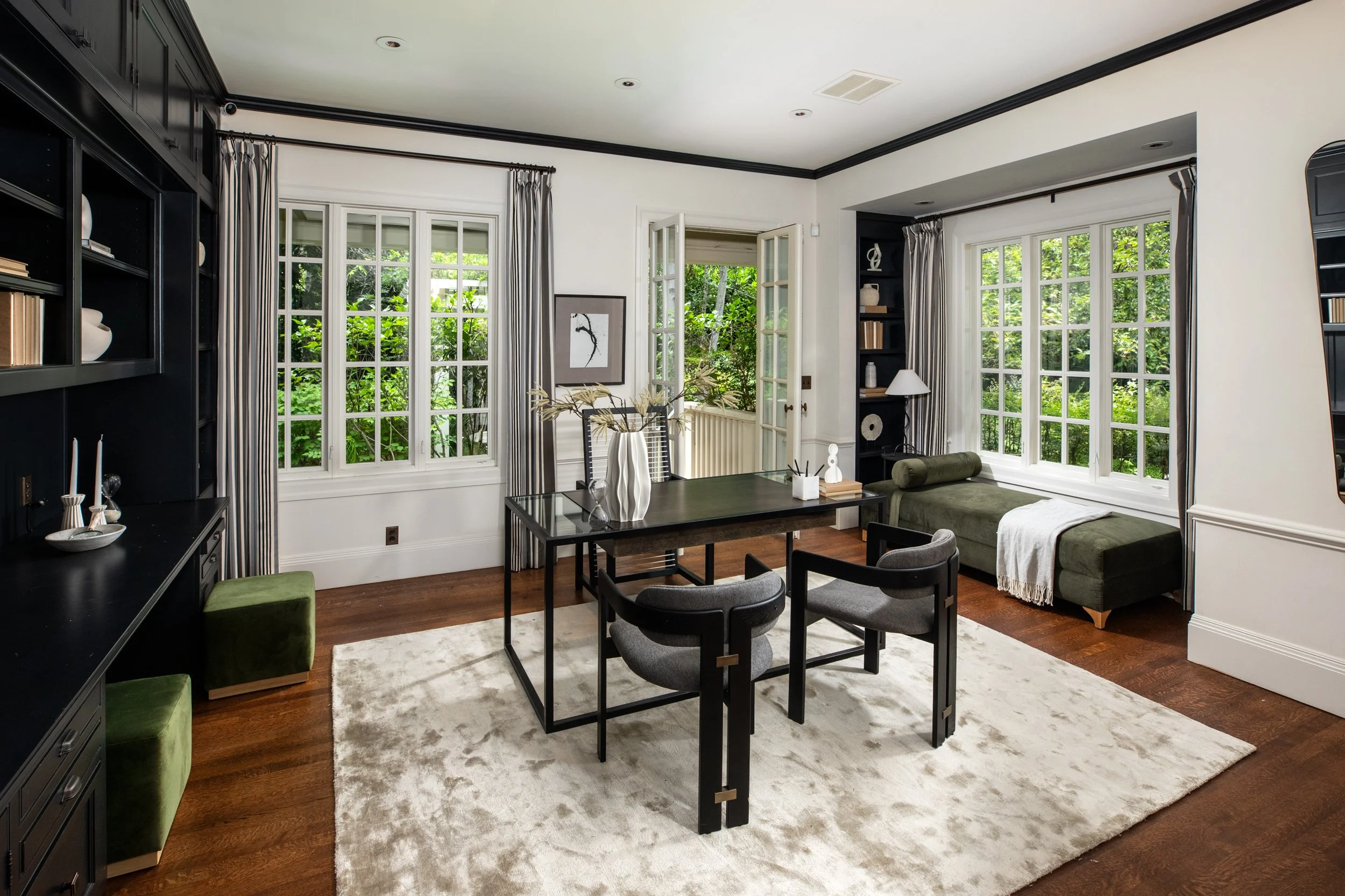 A bright living room with large windows showing greenery outside, black built-in shelves, a dining table with chairs, a beige rug, and a green daybed with a white throw blanket.