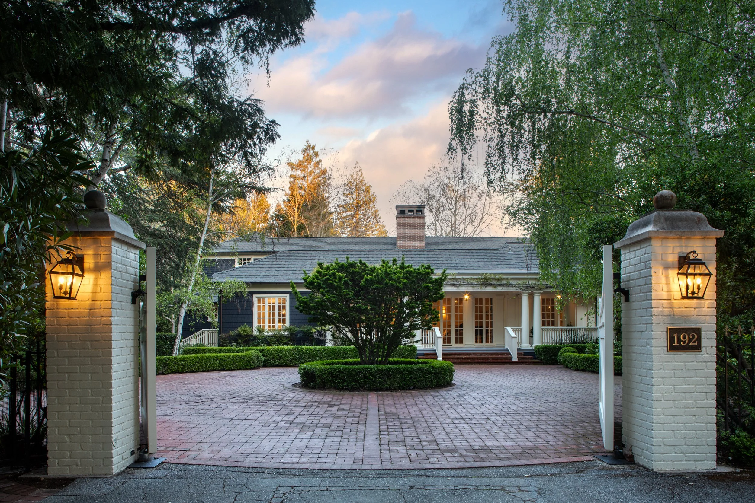 Front view of a house with a white porch, surrounded by landscaped garden and trees, viewed from an open gate with white brick pillars and lanterns.