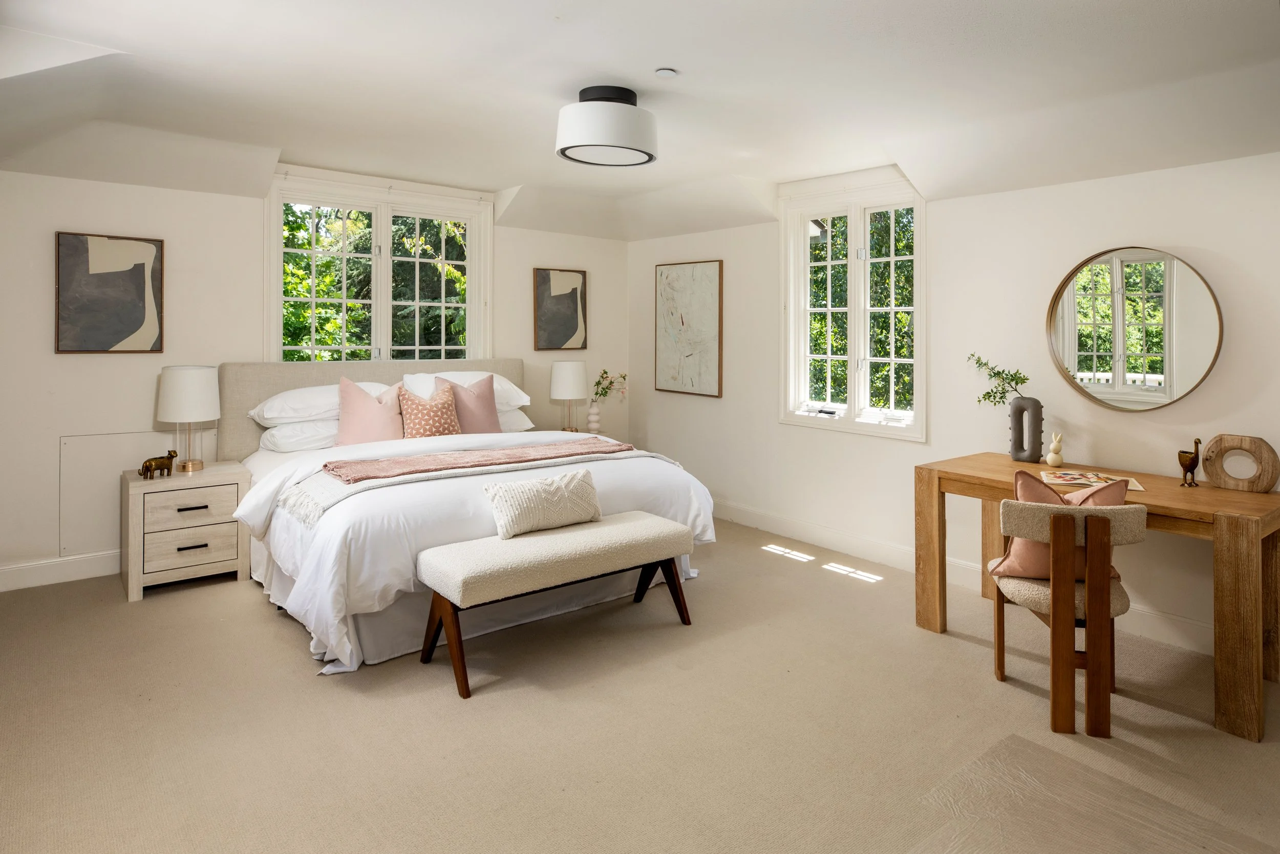 A cozy bedroom with a large bed, light-colored bedding, and decorative pillows, flanked by white nightstands with lamps, large windows with green foliage outside, and minimalist artwork on the walls. A wooden desk with decor and a mirror nearby.