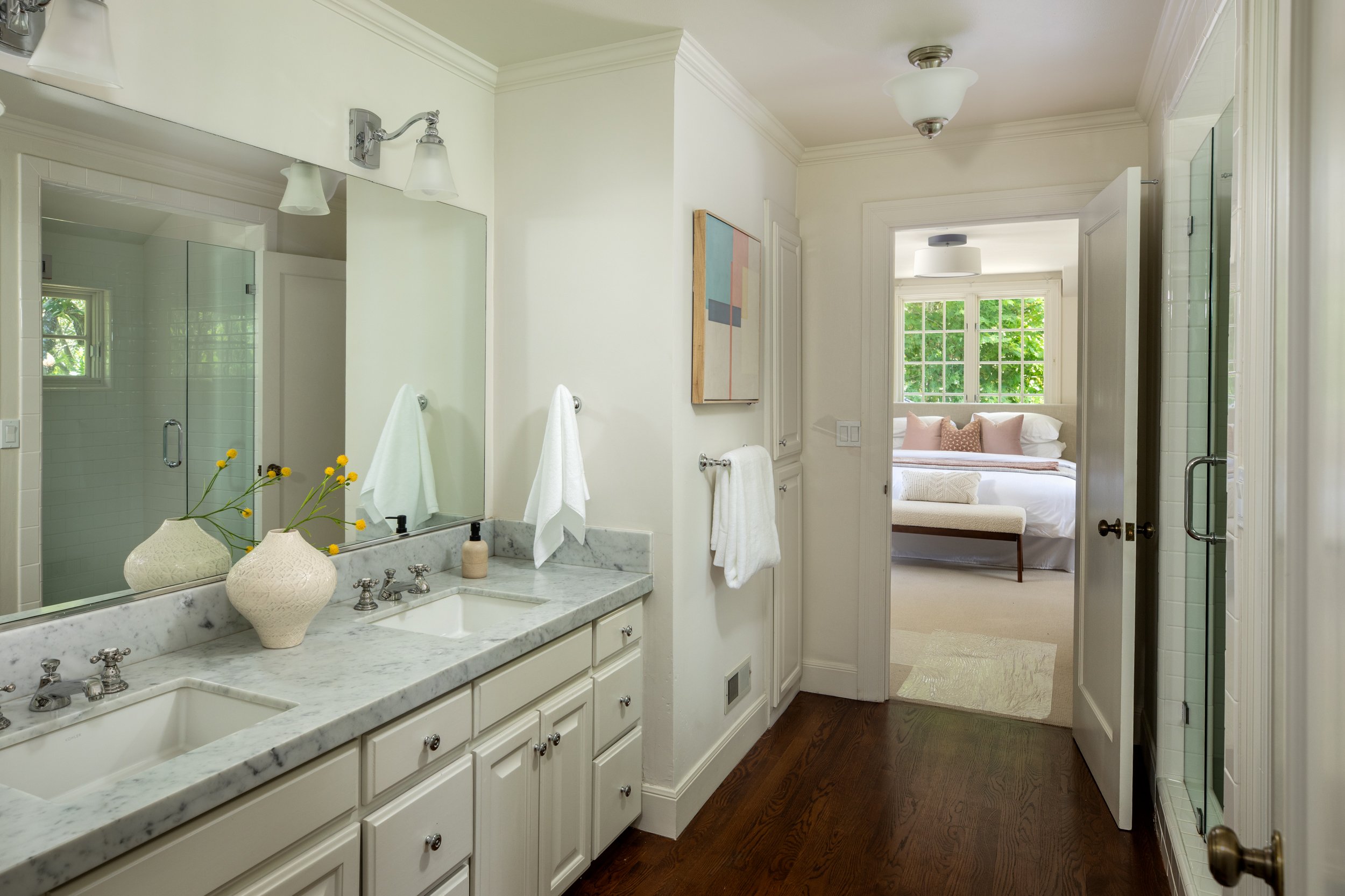 A bathroom with a marble countertop vanity with two sinks, large mirror, decorative vases, and white towels, connected to a bedroom with large windows, a bed with pillows, and a bench. The bathroom has white walls, built-in cupboards, and ceiling lig