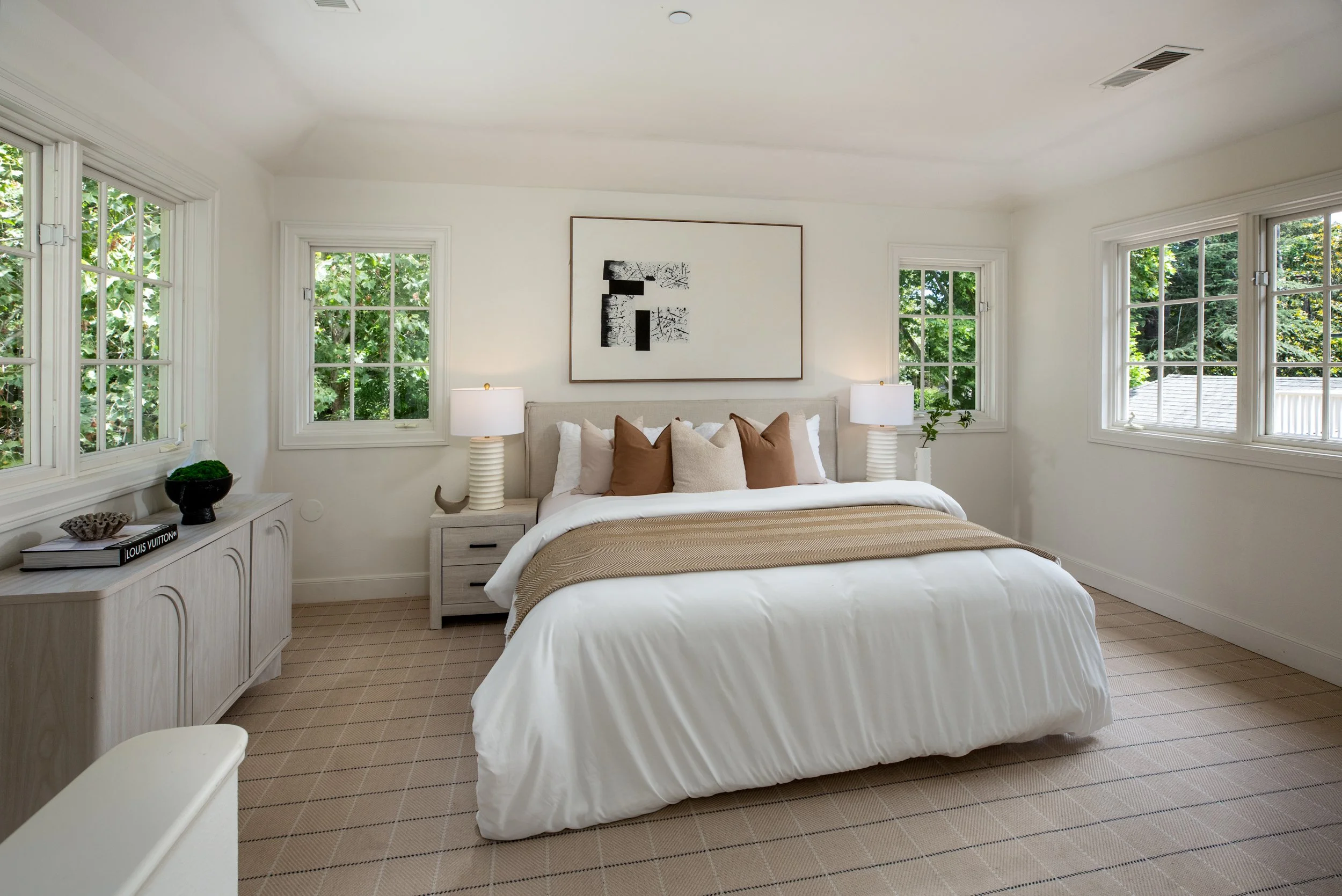 A cozy bedroom with a large bed covered in white bedding and beige pillows. There are two matching nightstands with white lamps on each side of the bed, and large windows with white frames providing a view of lush green trees outside. A piece of fram