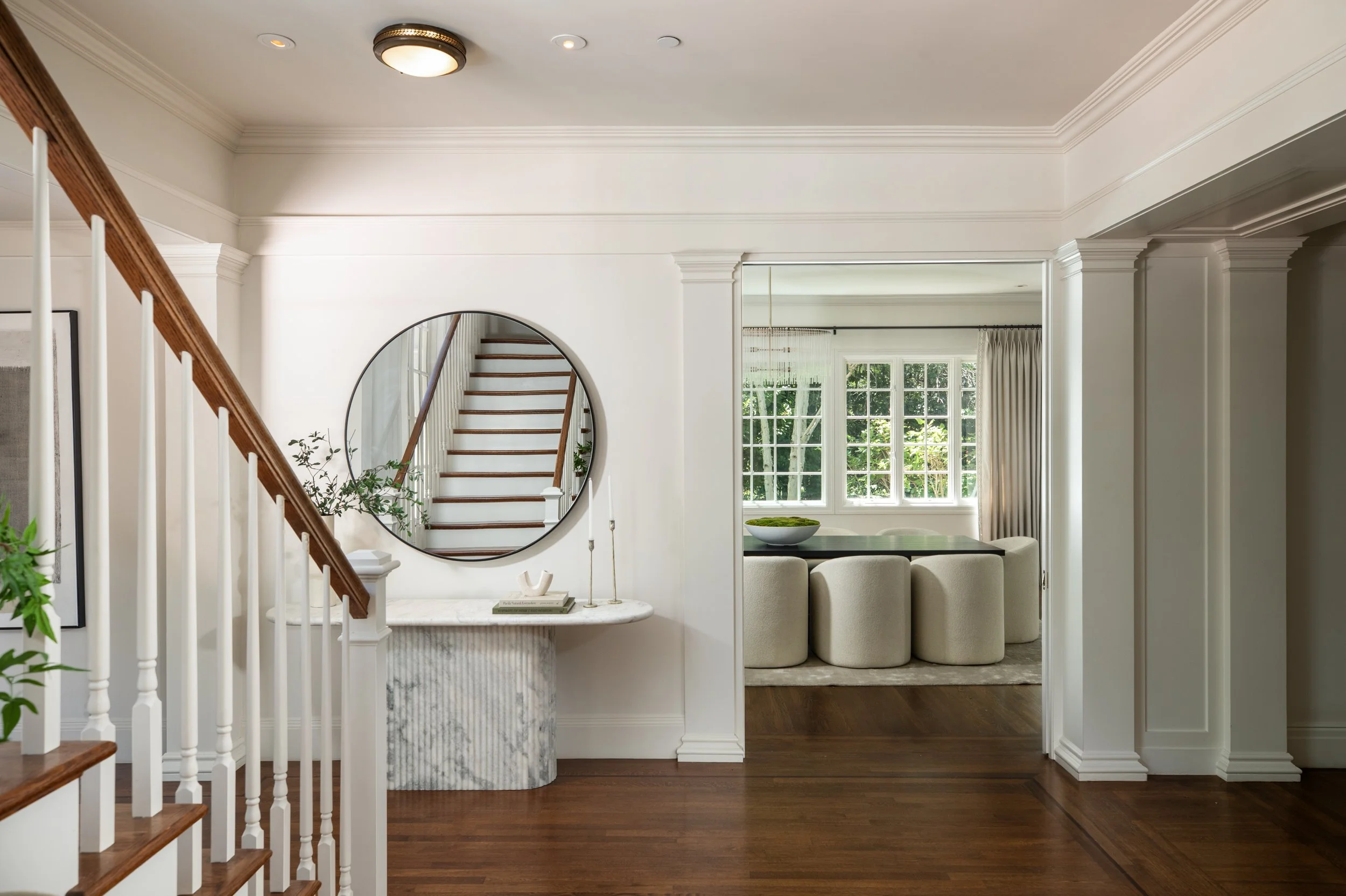 Interior view of a house with white walls, a staircase with wooden handrail, a round mirror on the wall, a marble-topped console table with decor items, a large window with curtains, and a separate room with a dining table and chairs in the backgroun