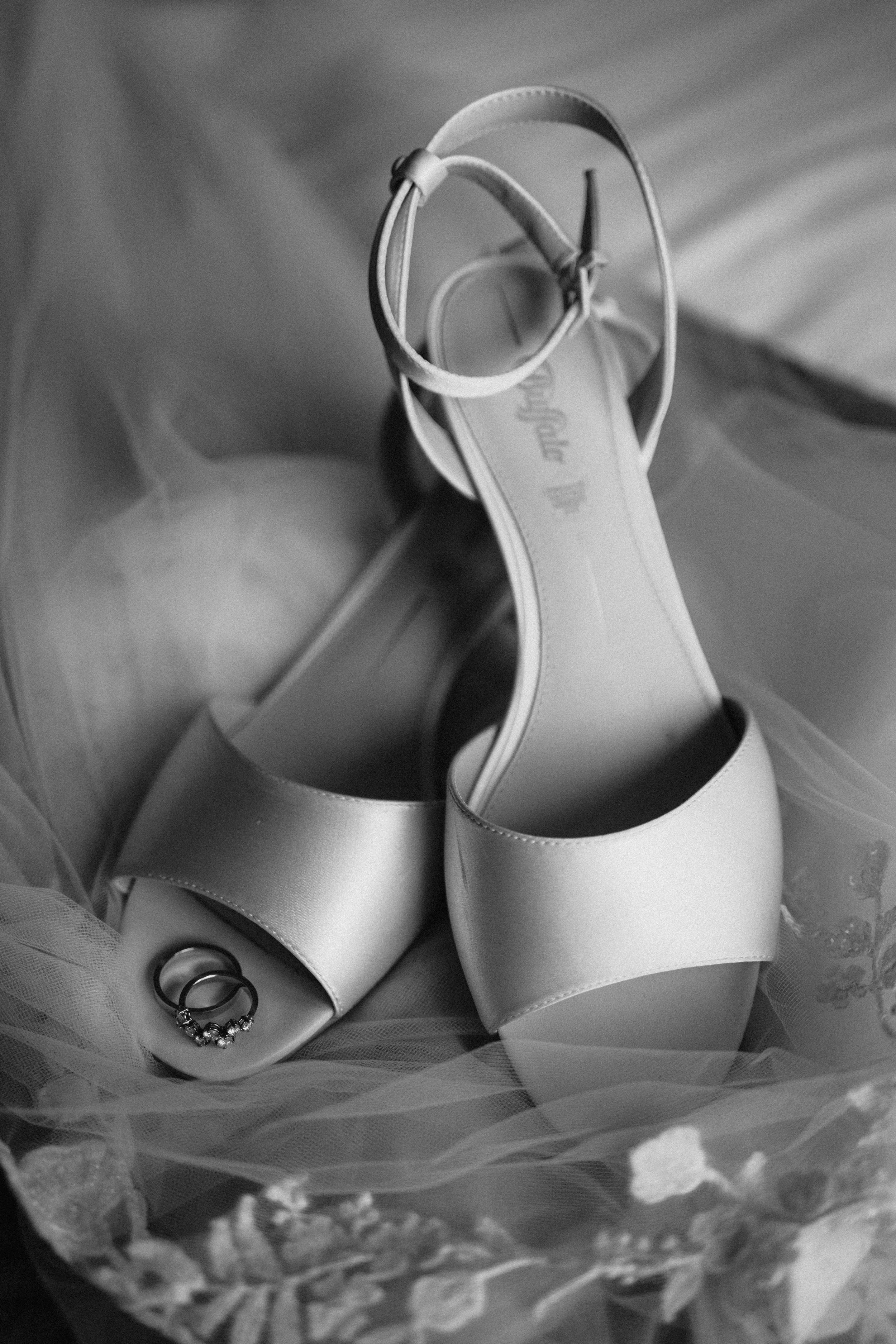 Zwart-wit foto van een paar elegante sandalen met hoge hakken en twee ringen, gelegen op een trouwsluier.