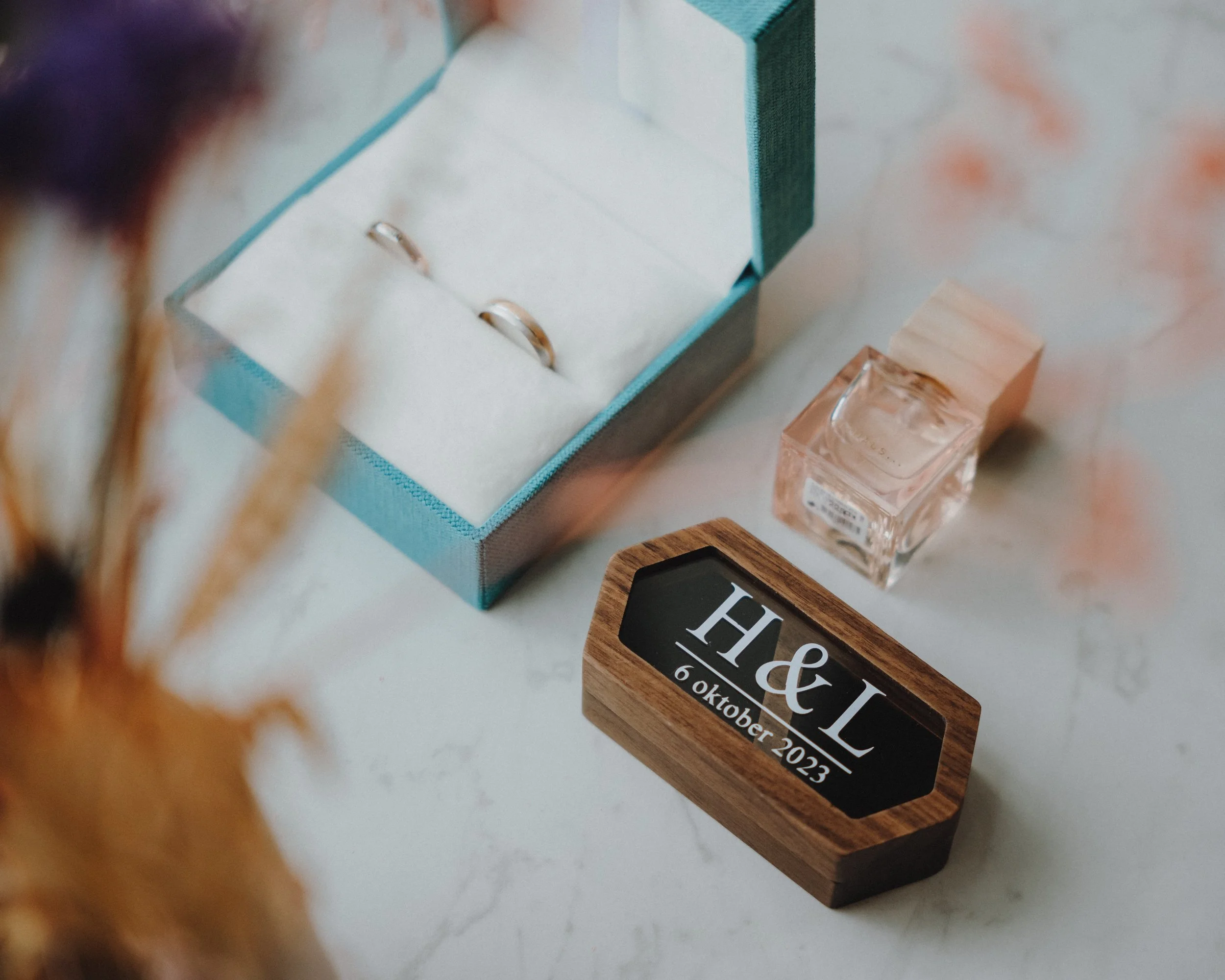 Twee ringen in een blauwe doos, een kleine fles parfum en een houten doosje met inscriptie "H&L 6 oktober 2023" op een marmeren oppervlak.