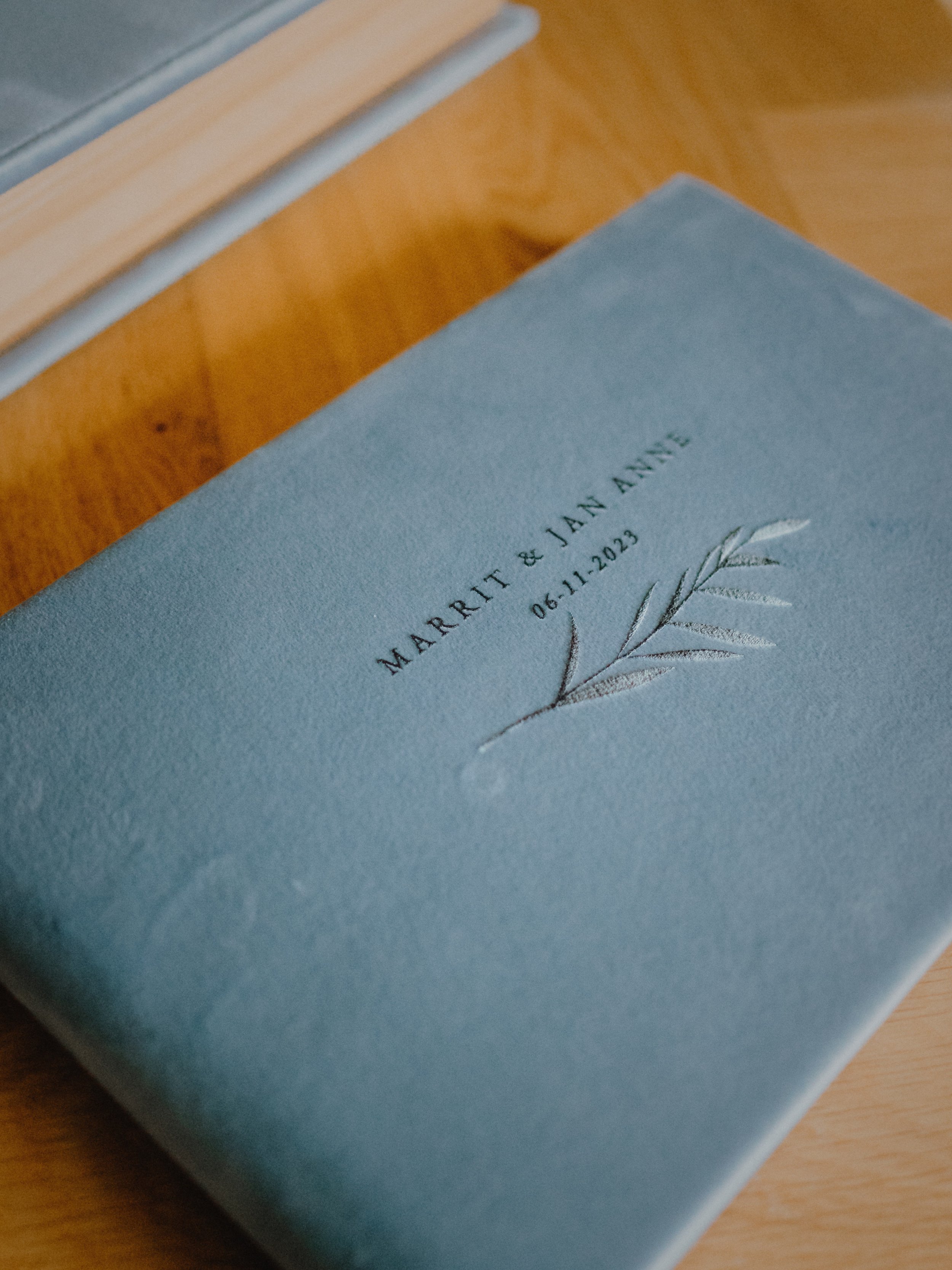 Een blauw trouwalbum met de namen "Marrit & Jan Anne" en de datum "06-11-2023" in een sierlijk lettertype op de kaft, met een subtiele decoratieve takkenmotief.