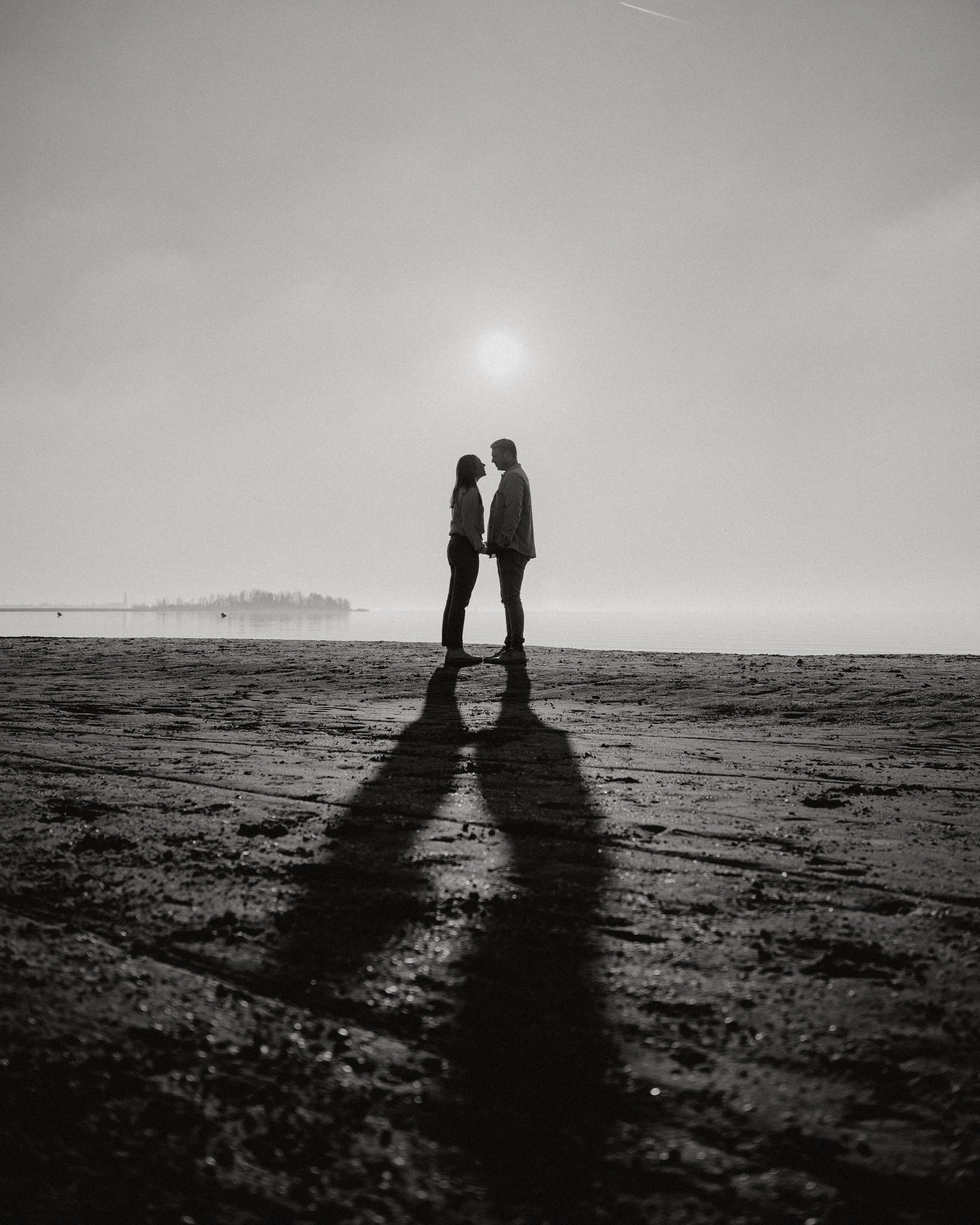 Zwart-wit foto van een paar dat elkaar vasthoudt op een strand bij zonsondergang, met lange schaduwen op de grond.