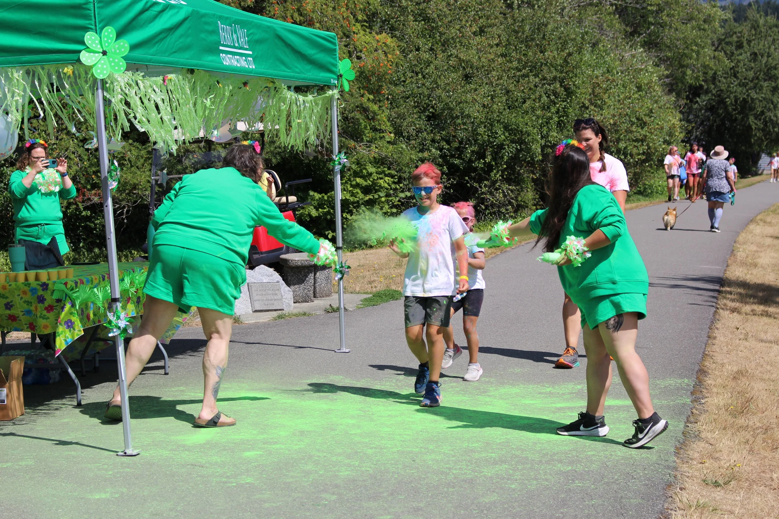 People running through a green flour station during a St. Patrick's Day themed fun run or parade, with onlookers and dog walkers in the background.