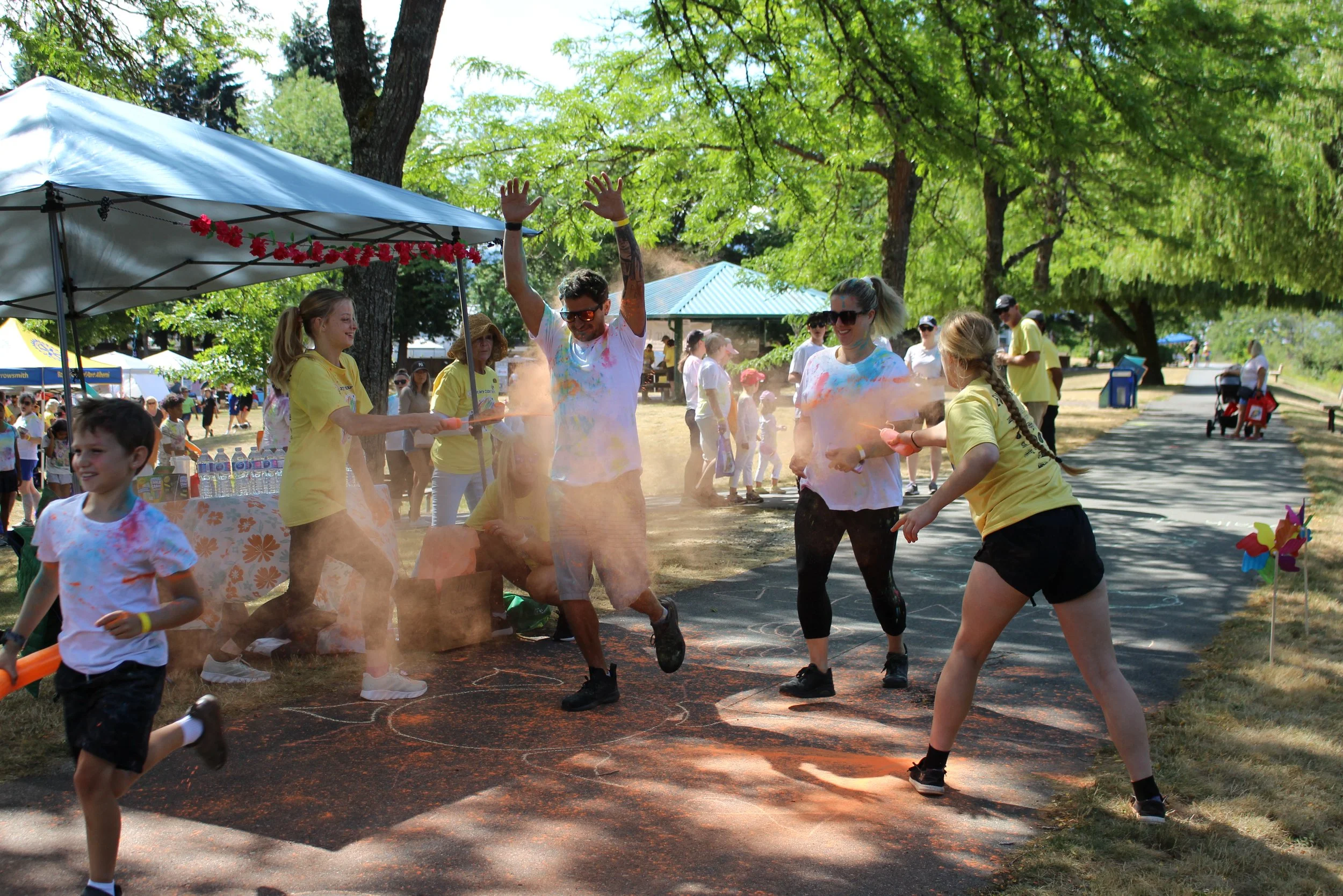 People participating in a color powder run event, throwing colored powder at each other on a sunny day in a park with trees, tents, and other participants in the background.