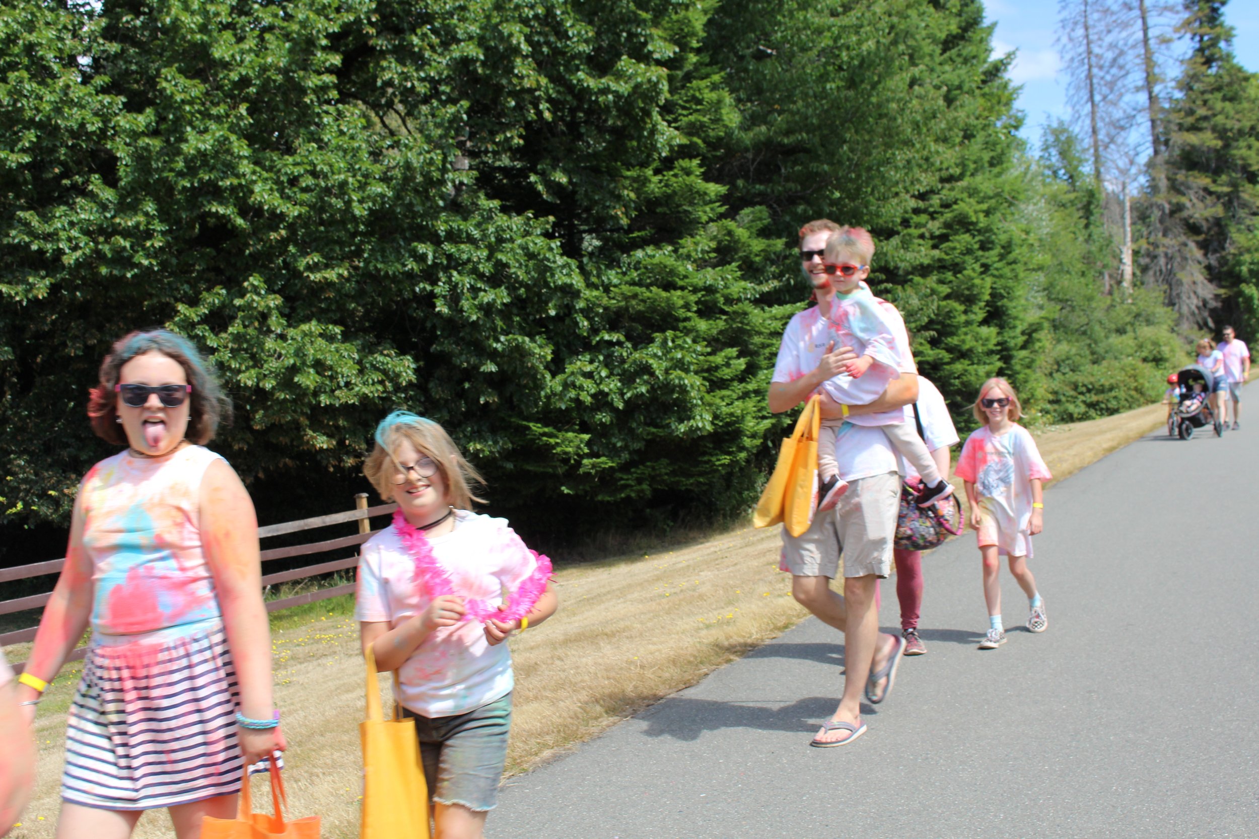 Group of children and adults walking outdoors on a sunny day, some smiling and wearing colorful clothes, near a wooded area.