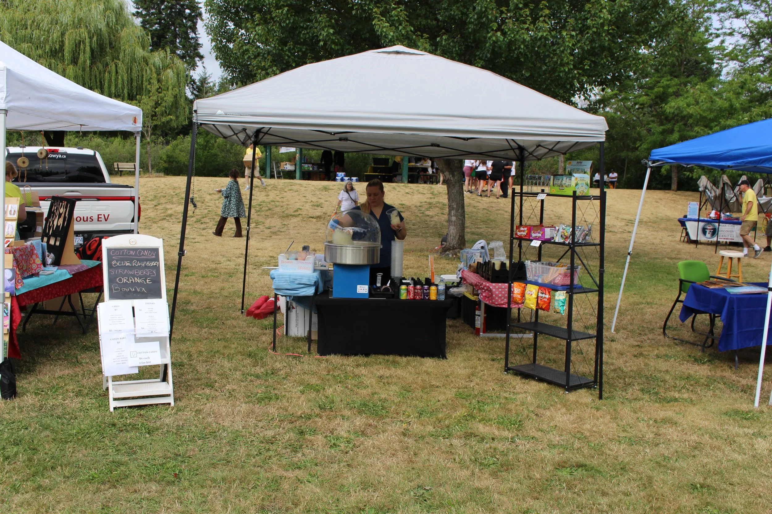 A food stand at an outdoor market, with a woman serving cotton candy, and various snacks and items on display, set up under a white canopy on grassy ground.
