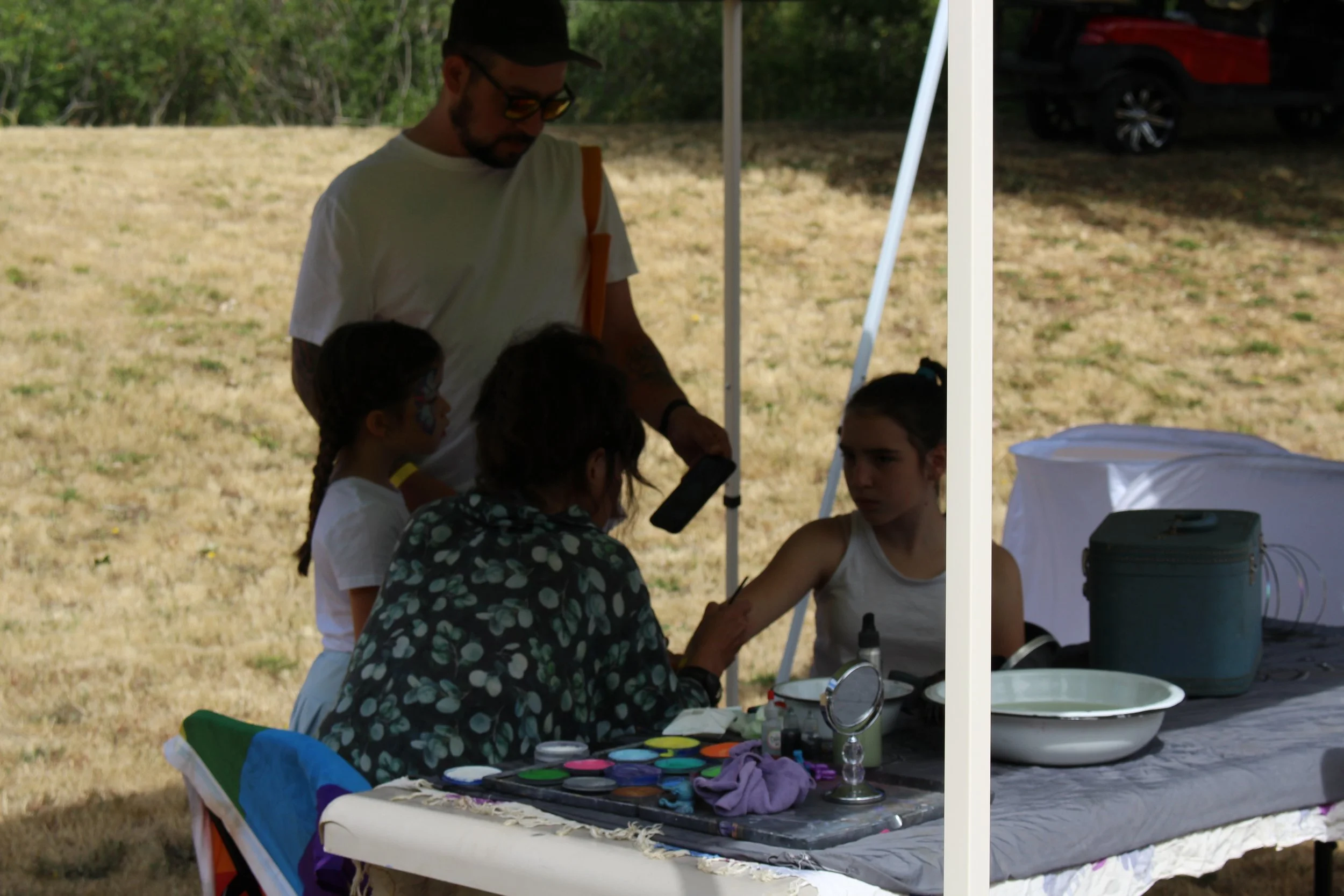 A face painting activity taking place outdoors under a white tent in a park or grassy area, with children and an adult engaged in the activity.