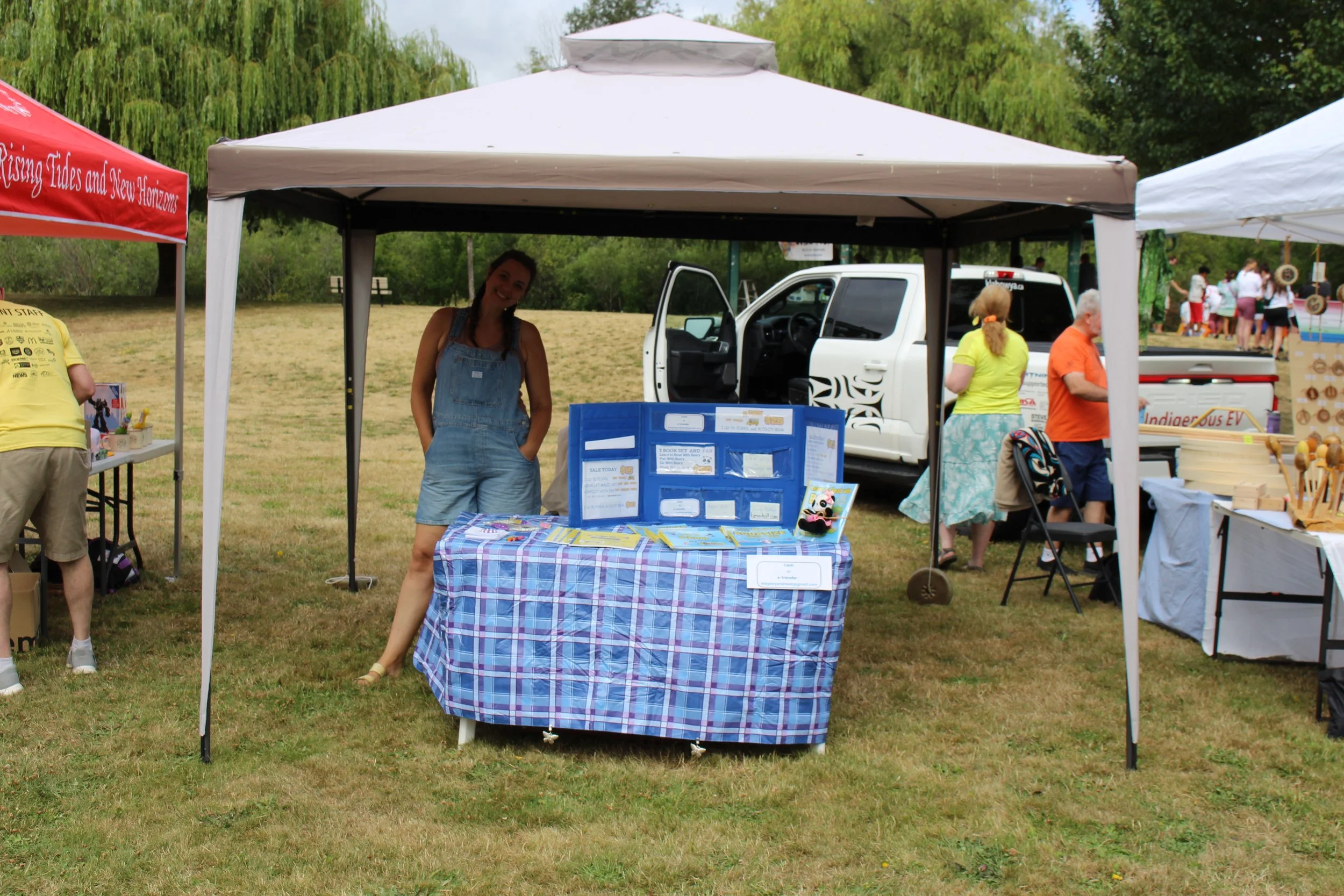 A woman standing behind a blue plaid table under a white canopy at an outdoor event. The table has informational materials and a display board. Several other tents and people are visible in the background.