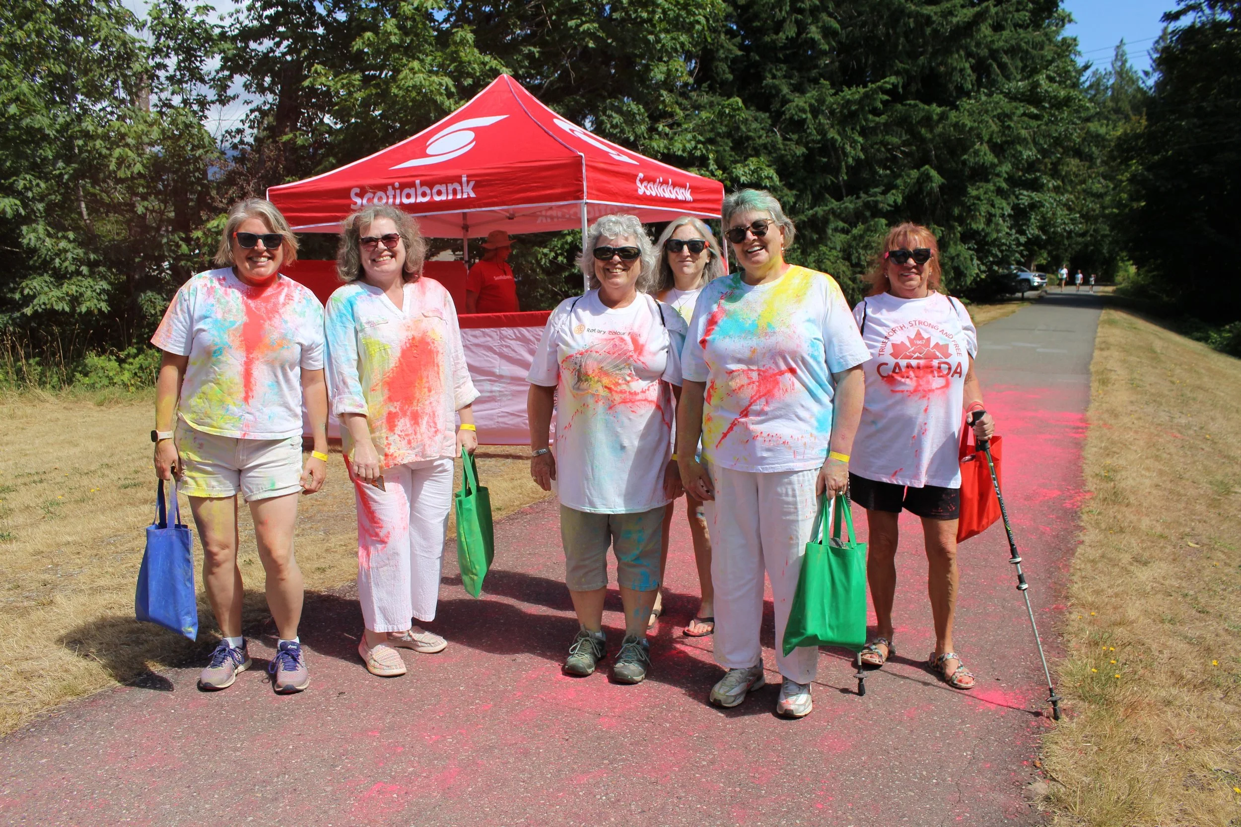 Group of women smiling and covered in colorful powder, standing outdoors on a pink trail during a color run event. They are wearing white T-shirts and holding green bags, with a red canopy tent in the background under a clear sky and surrounded by tr