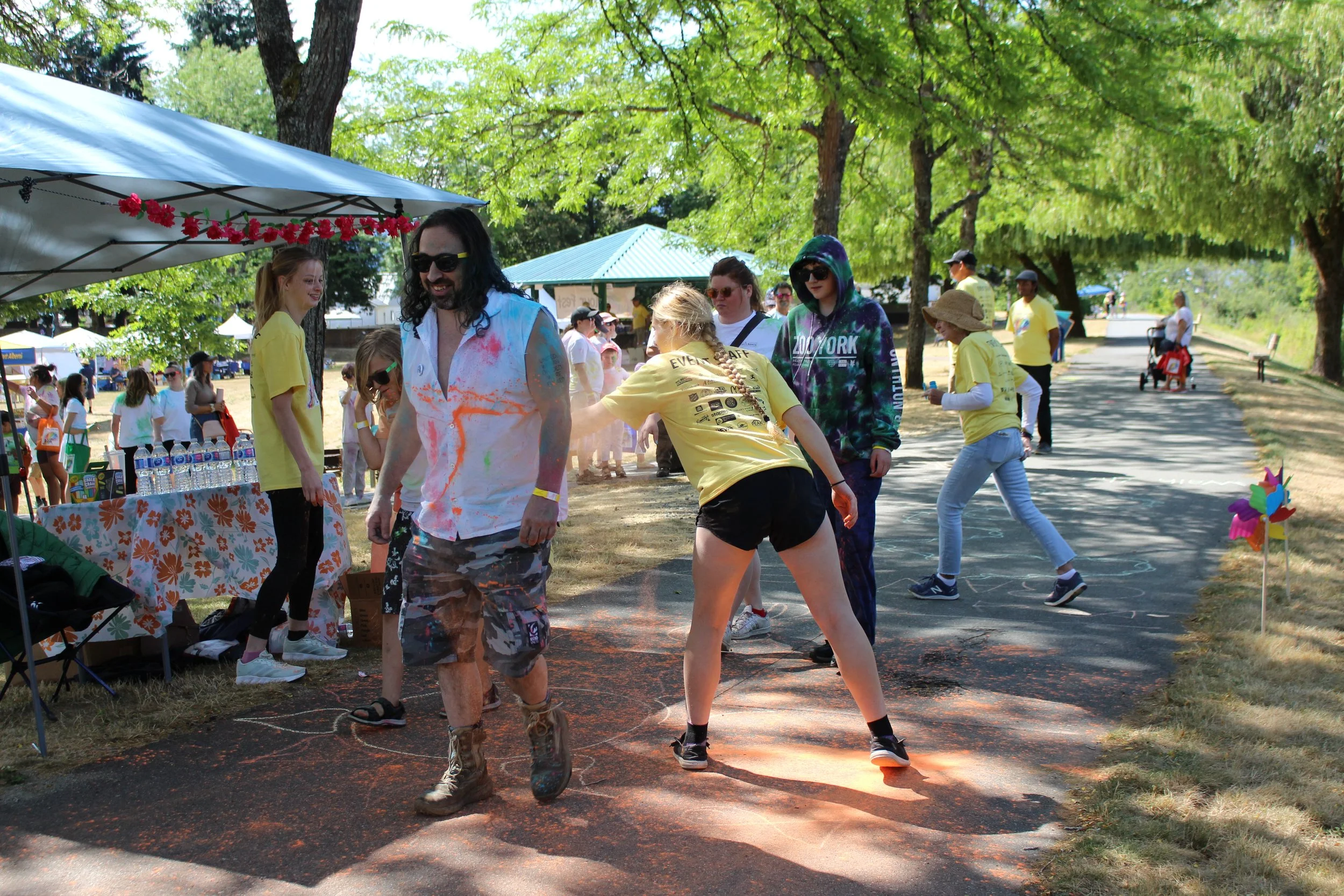 People participating in a color run event outdoors on a sunny day, with trees and a park trail in the background. Some are covered in colored powder, and there are tents and onlookers nearby.
