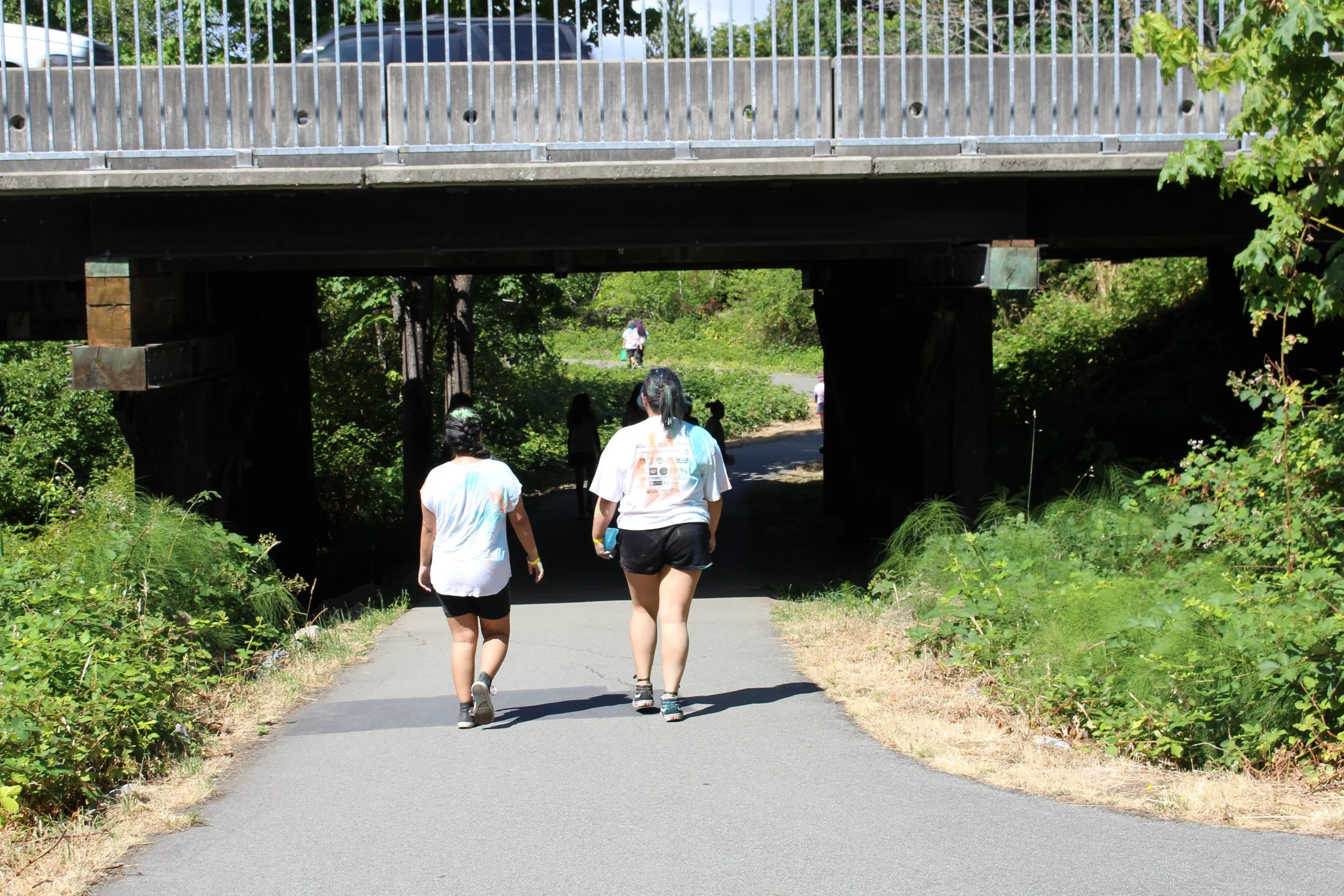 People walking on a paved trail under a bridge in a green, wooded area.