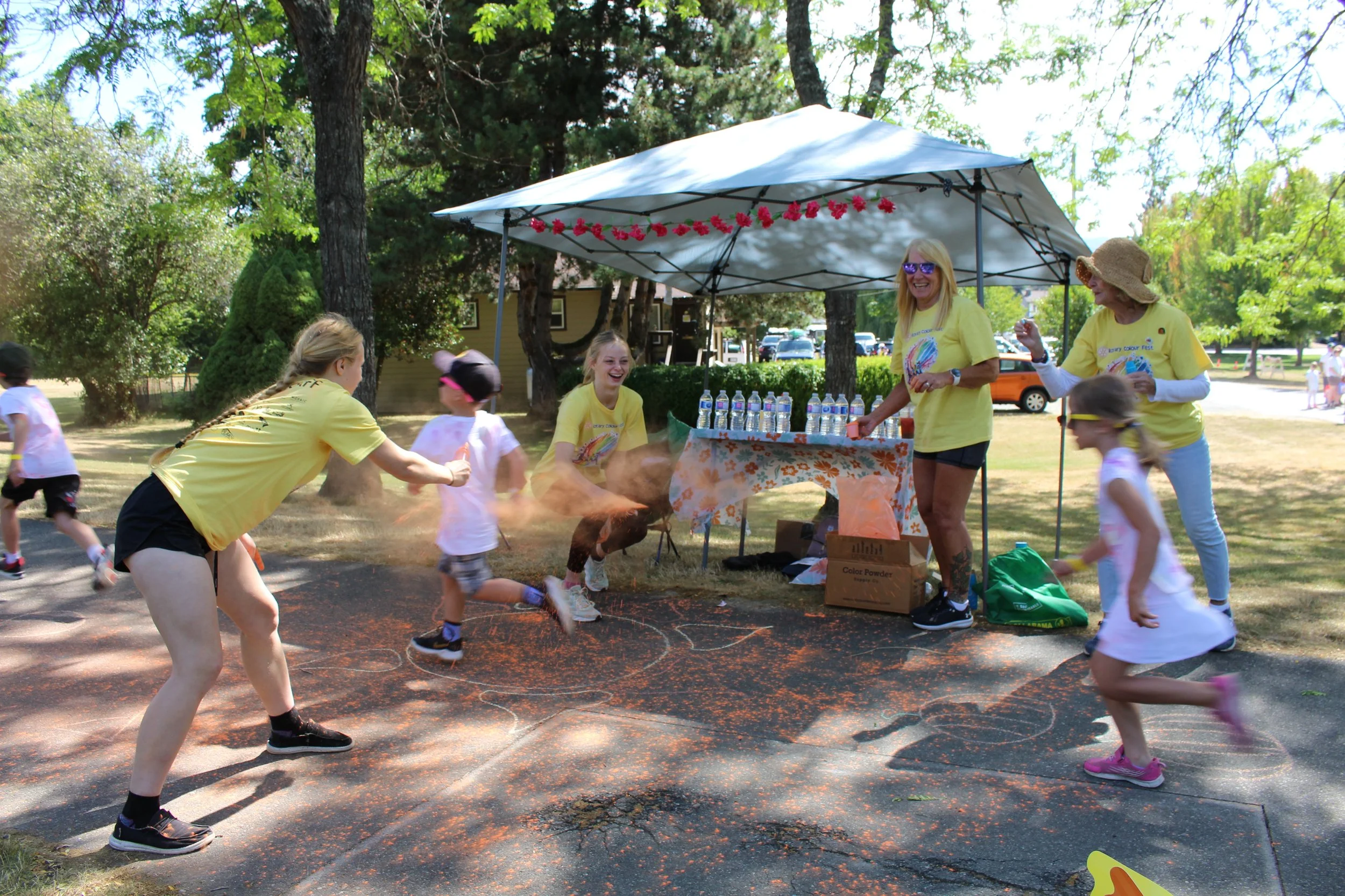 Children and adults engaging in a color powder fight around a booth with water bottles on top, outdoors in a park with trees and a sunny sky.