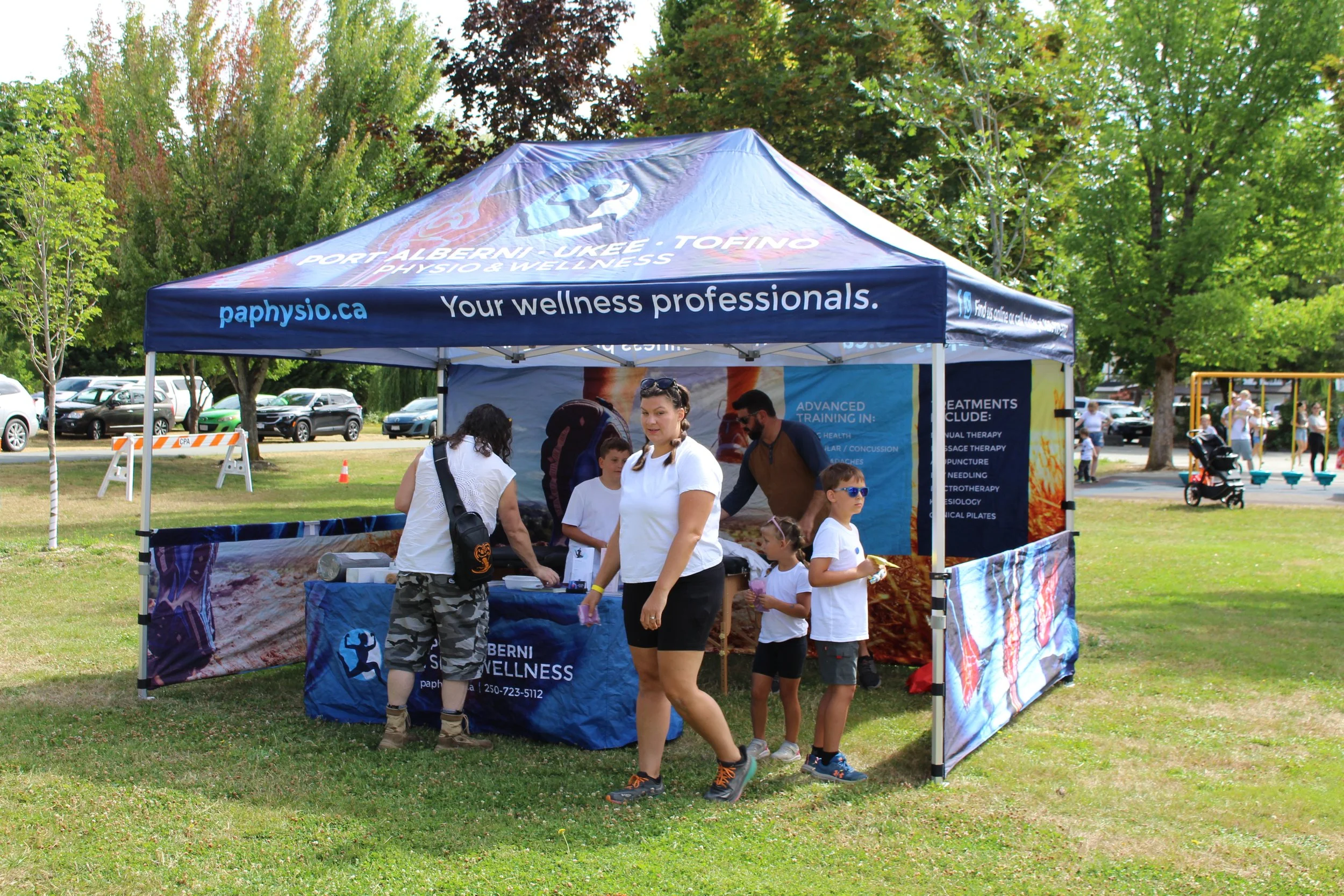 A group of people gathered at a wellness booth set up in a park with a blue canopy tent labeled 'Port Alberni, Ukee, Tofino - Physio & Wellness'. The booth displays informational posters about wellness services. Some children and adults are engaging 